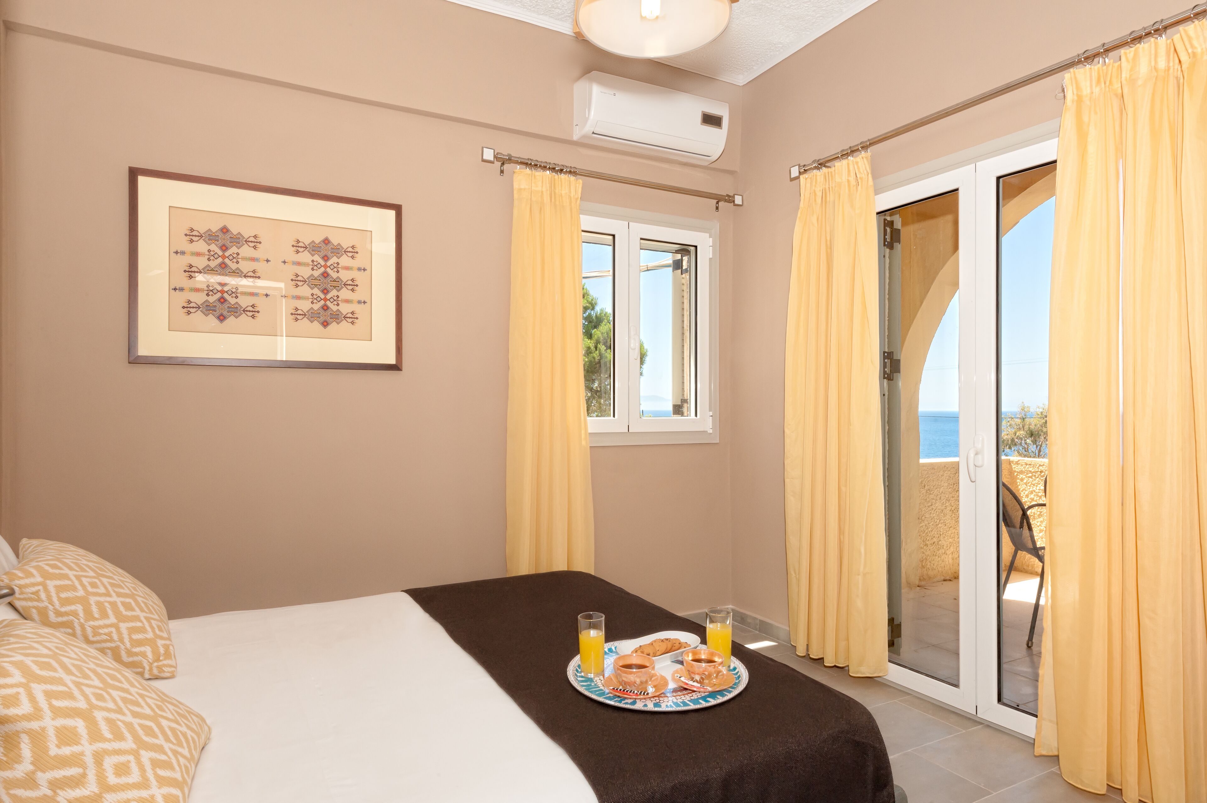 Ein Schlafzimmer mit Bett, einem Frühstückswagen mit Gebäck und Orangensaft, einem gerahmten Bild an der Wand und Meerblick durch eine Schiebetür.