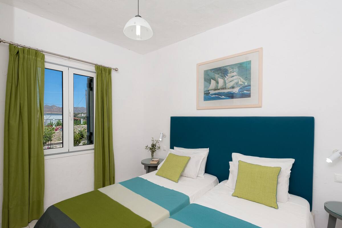 A hotel room with two beds, green curtains, a painting of a sailboat, and a view of the outdoors.