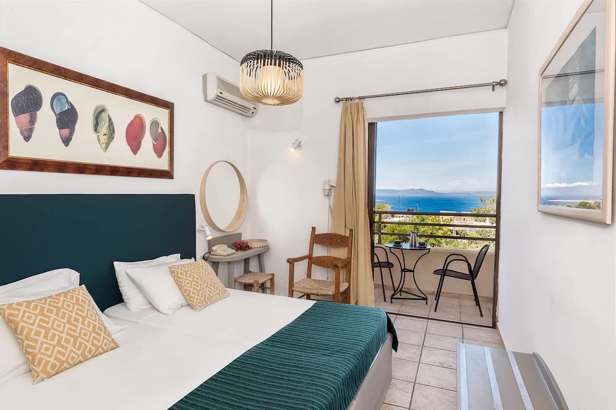 A hotel room with a bed, a chair, a table, a balcony with a view of the sea, and a framed picture on the wall.