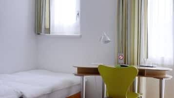A small, neatly arranged room with a single bed, a desk, a chair, and a window with curtains.