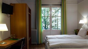 Una habitación de hotel con una cama, un escritorio con lámpara, una silla, un radiador y una ventana con cortinas.