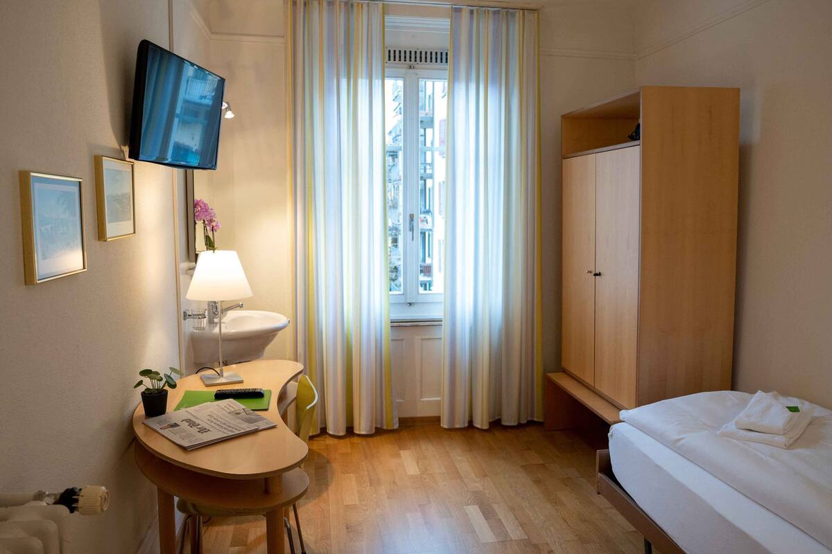 Ein Hotelzimmer mit einem Bett, einem Schreibtisch mit Lampe, einem Fernseher und einem Fenster mit Vorhängen.