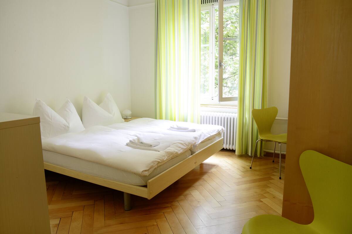 Ein ordentlich bezogenes Bett mit weißen Leinen und Kissen, ein leuchtend grüner Sessel und ein Fenster mit grünen Vorhängen.