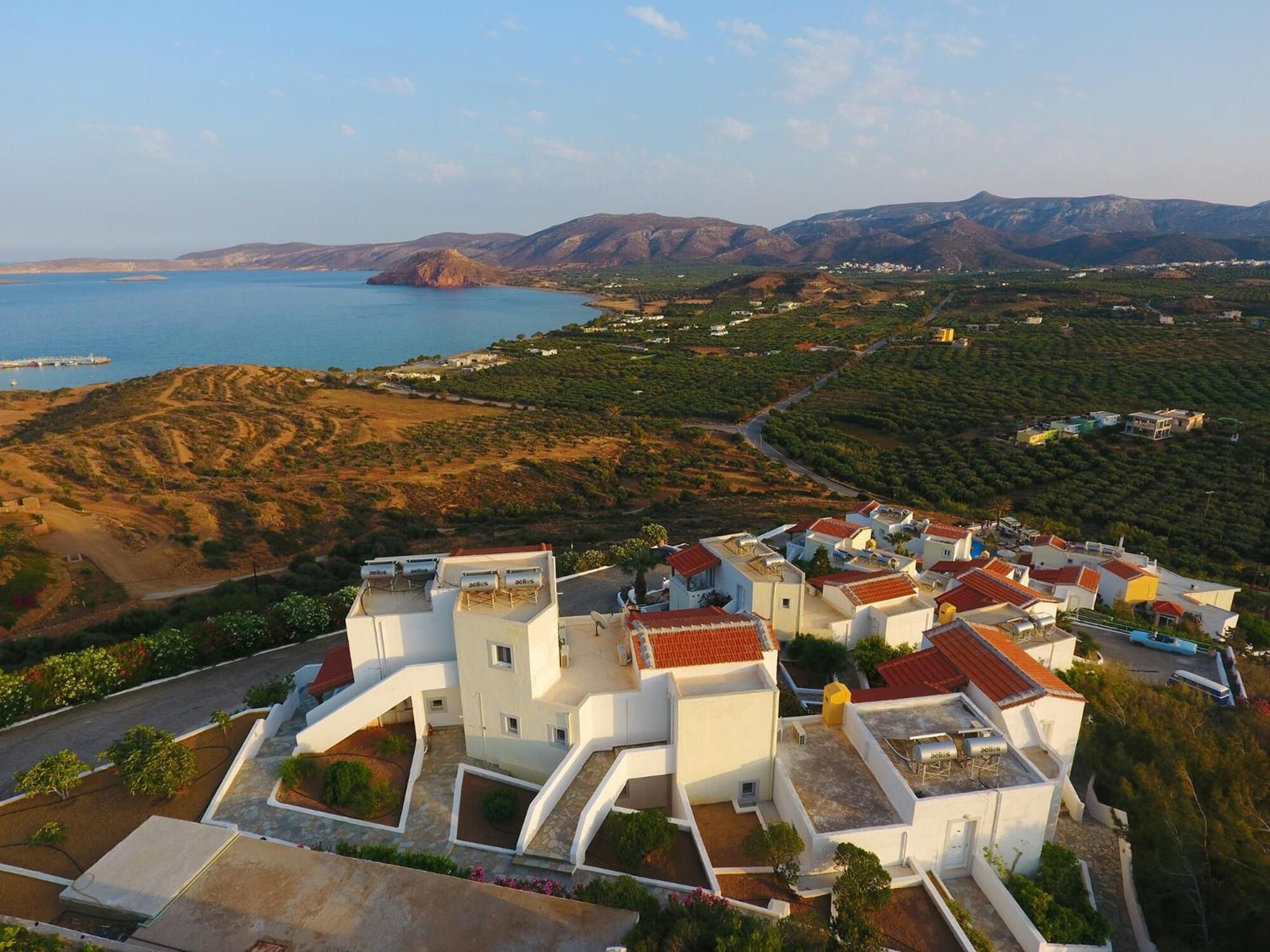 A coastal village with white buildings and red roofs, surrounded by hills and a body of water.