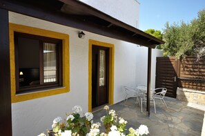 A white building with yellow trim around windows and doors, a stone patio with metal chairs, and a wooden gate. - Marina Village (Sitia)