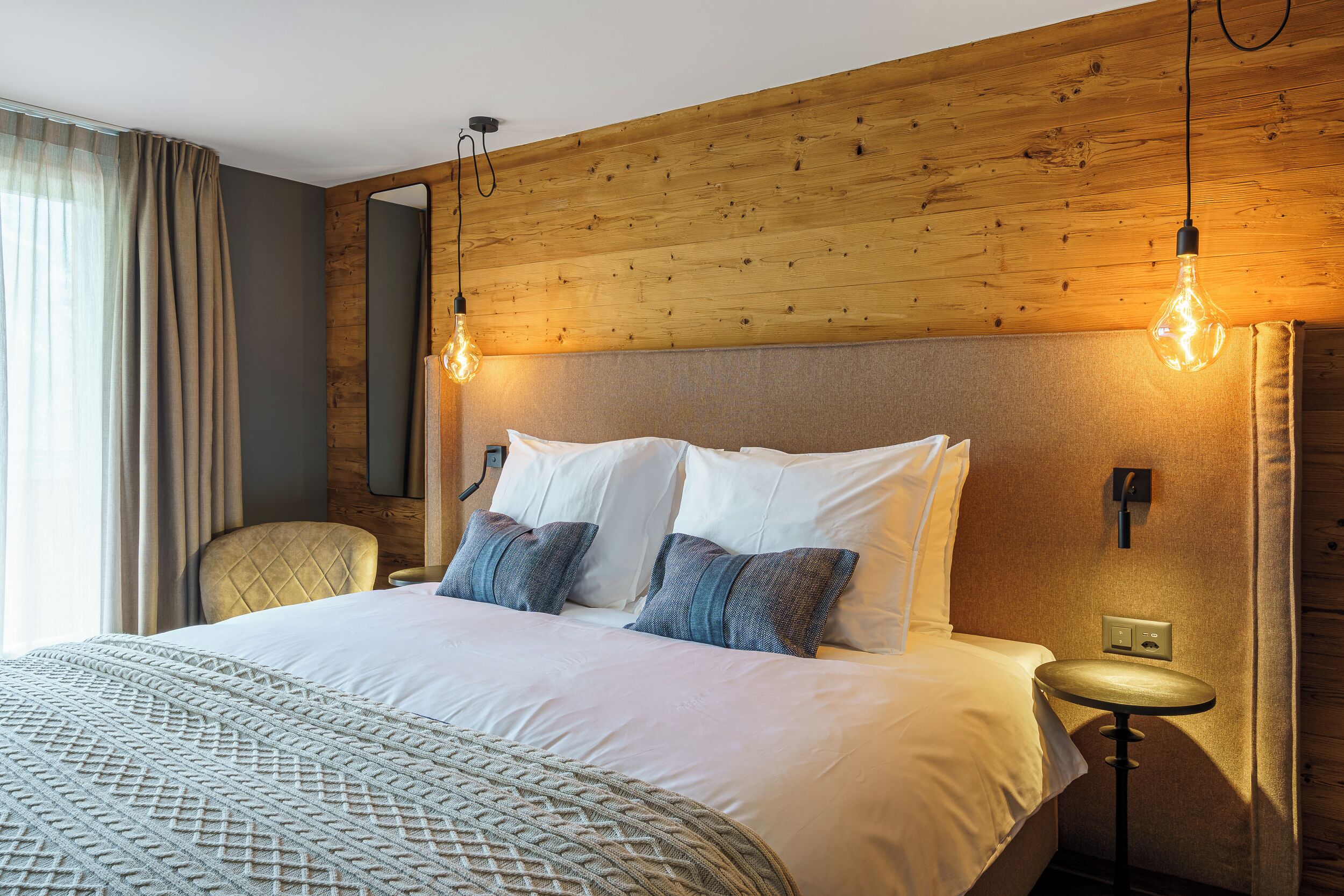 Ein Hotelzimmer mit hölzernem Kopfteil, einem Bett mit weißer und grauer Bettwäsche, einem runden Beistelltisch und einem Sessel.