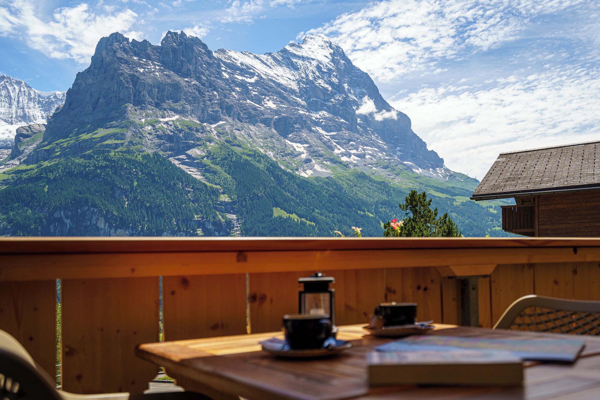 A wooden table with a view of a mountain range, a wooden cabin, and a clear sky.