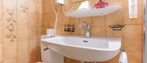 A bathroom with a white sink, a toilet, and a shower area. There is a hairdryer on the wall, a vase with a flower, and a bottle of lotion on the shelf.