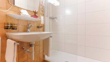 Ein Badezimmer mit Waschbecken, Spiegel und Dusche. Handtücher hängen an der Wand und ein Ablagebereich enthält Toilettenartikel.