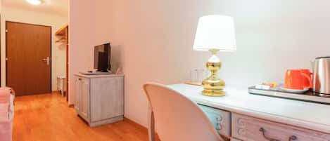 Una habitación con piso de madera, un escritorio con lámpara, una silla, un televisor y una puerta.