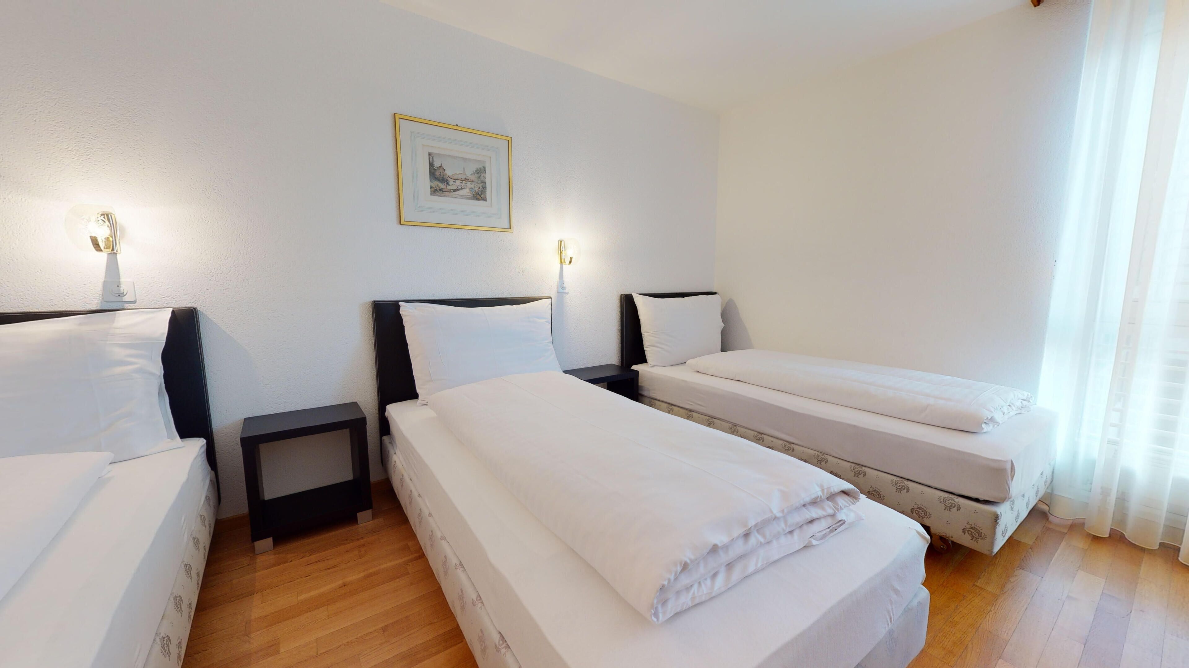 Ein Hotelzimmer mit zwei Einzelbetten, einem kleinen Nachttisch mit Lampe, einem gerahmten Bild an der Wand und einem Fenster mit durchsichtigen Vorhängen.
