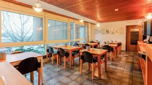 A dining area with wooden tables and chairs, large windows with a view of trees, and a wooden ceiling. - Hôtel du Grand-Pré (Marly)