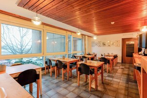 Un comedor con mesas y sillas de madera, ventanas amplias con vistas a árboles y techo de madera.