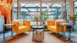 A bright room with yellow sofas, a glass table, and a view of a green structure outside. - Hôtel du Grand-Pré (Marly)