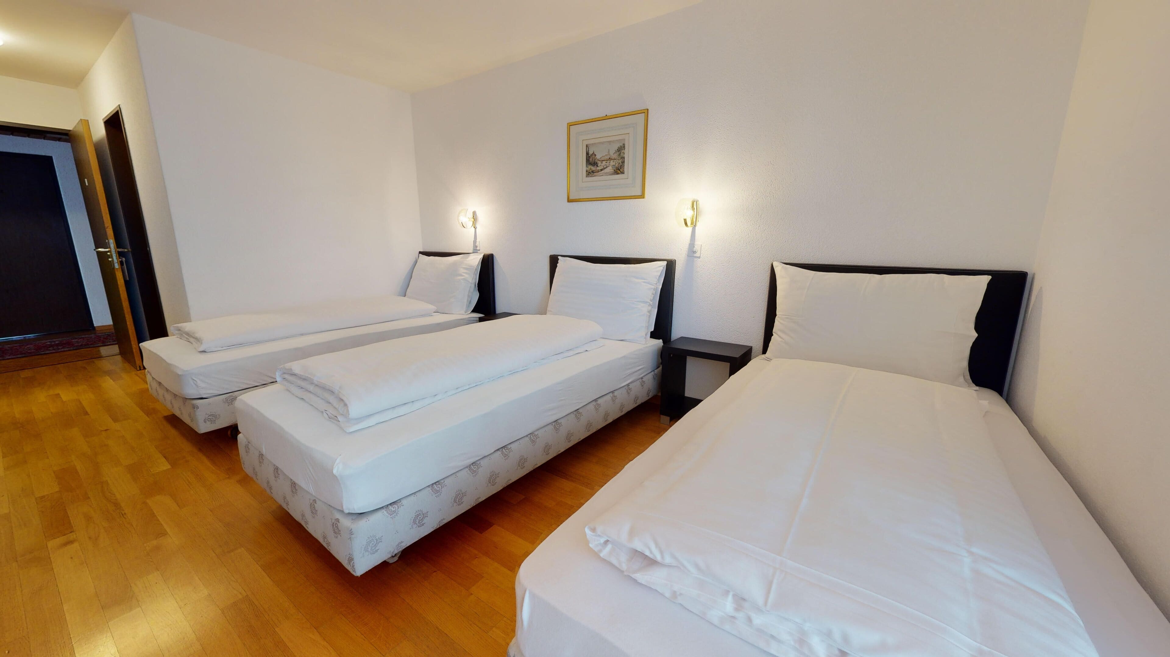 A hotel room with three single beds, wooden flooring, and a framed picture on the wall.