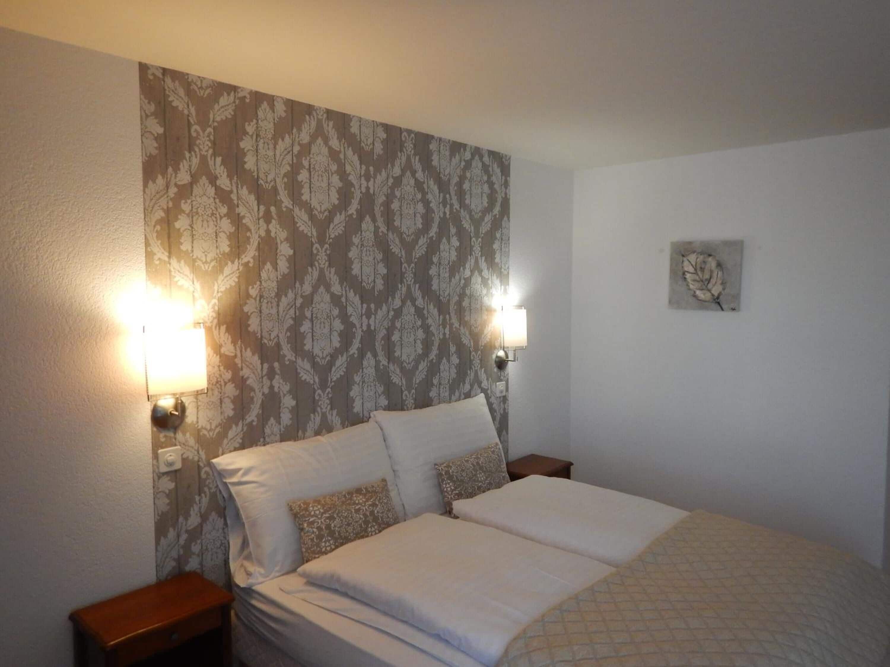 Una cama bien hecha con dos almohadas, una pared con papel tapiz estampado y dos lámparas de pared.