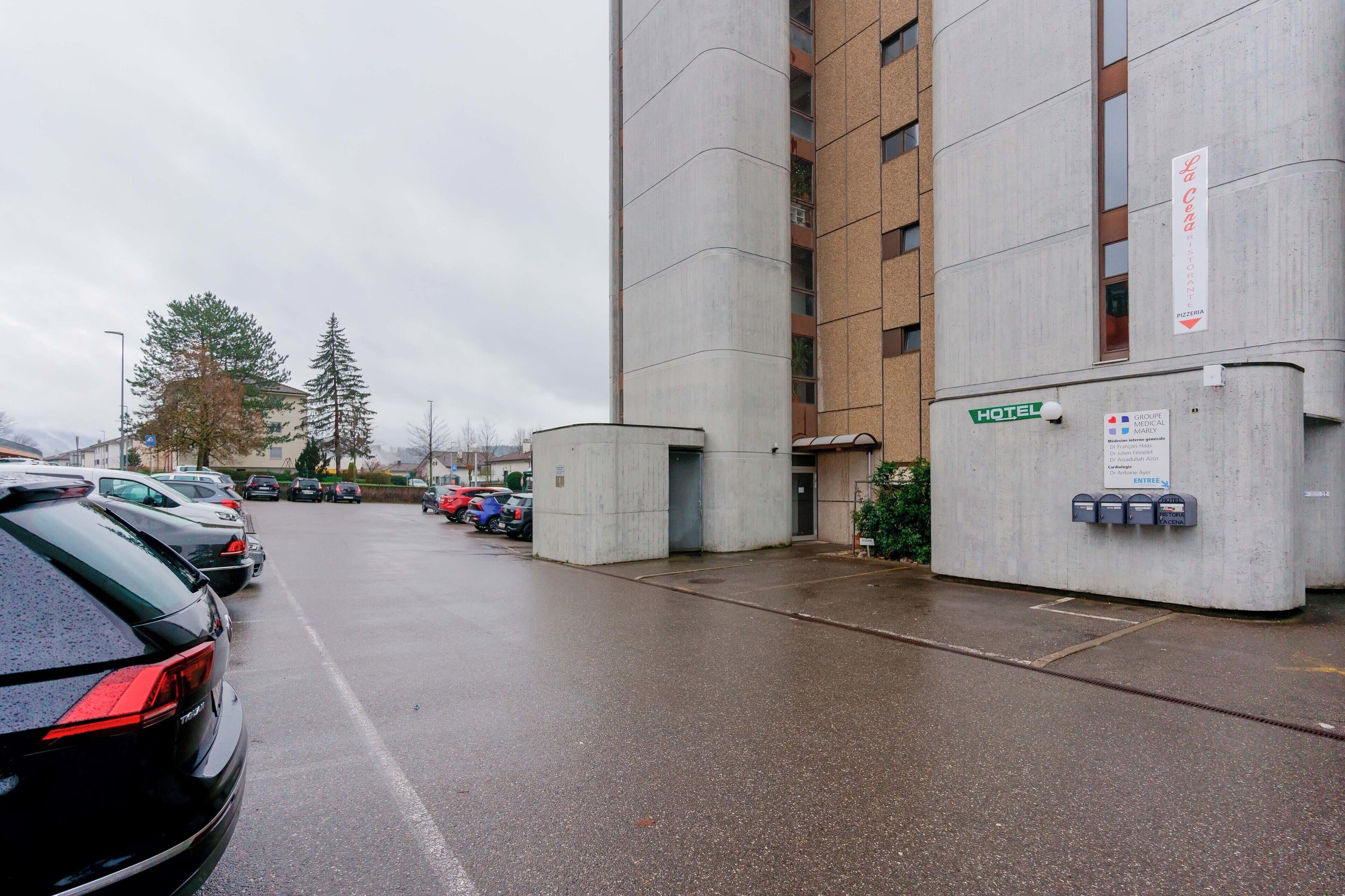 A hotel entrance with a concrete facade, a sign indicating a medical group, and a parking lot with cars.