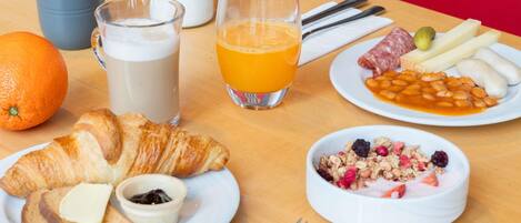 A breakfast table with croissants, a bowl of cereal, a plate of beans, a glass of orange juice, a glass of milk, and a small potted plant.