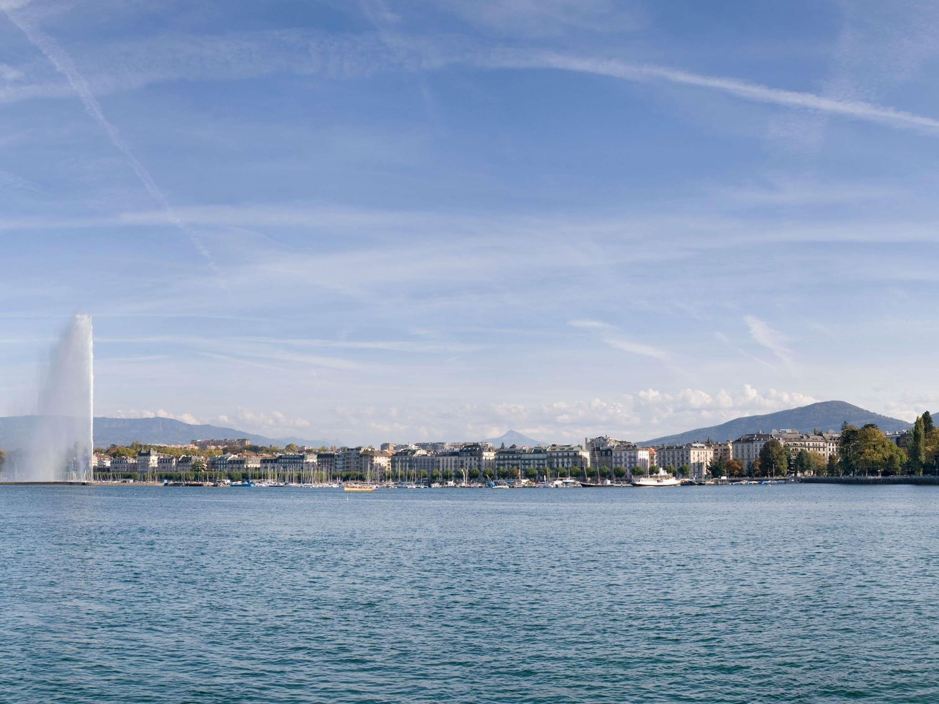 Un paisaje urbano frente al agua con una fuente destacada, edificios y una montaña al fondo.