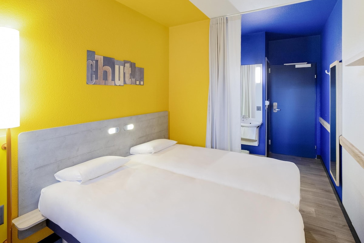 Quarto de hotel com cama de casal, mesa de cabeceira, espelho e banheiro visível por uma porta.