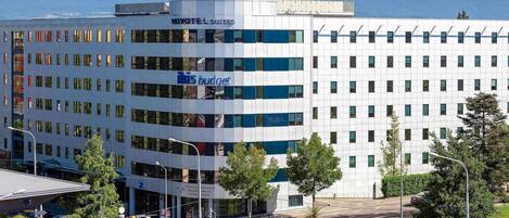 A modern hotel with a prominent 'Novotel Suites' sign, a 'His Budget' restaurant, and a view of mountains in the background.