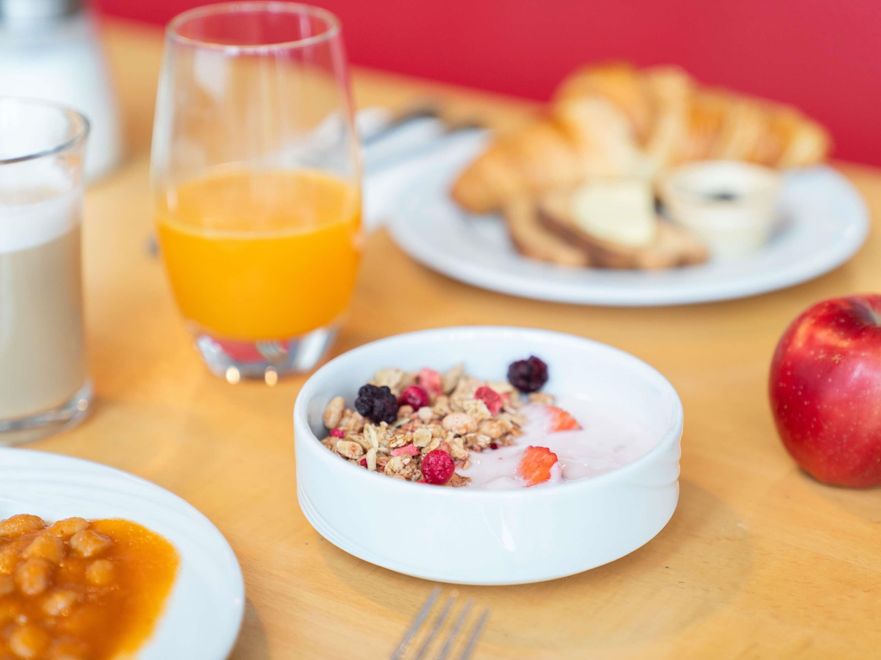A breakfast setting with a bowl of cereal, a glass of orange juice, a croissant, and a red apple on a wooden table.