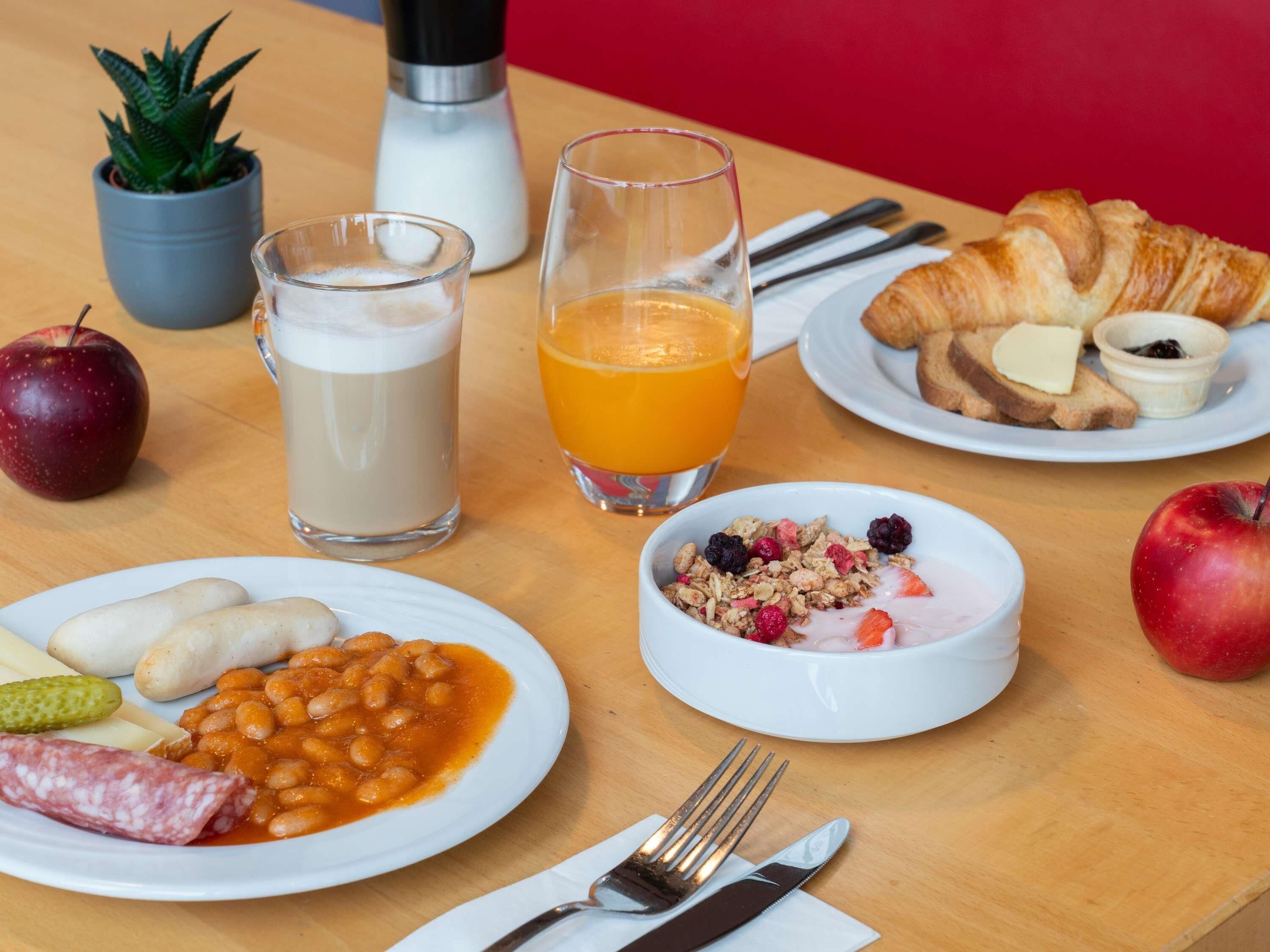 Una mesa de desayuno con platos de frijoles, salchichas, tostadas, cruasanes y fruta. También hay vasos de leche y jugo de naranja.