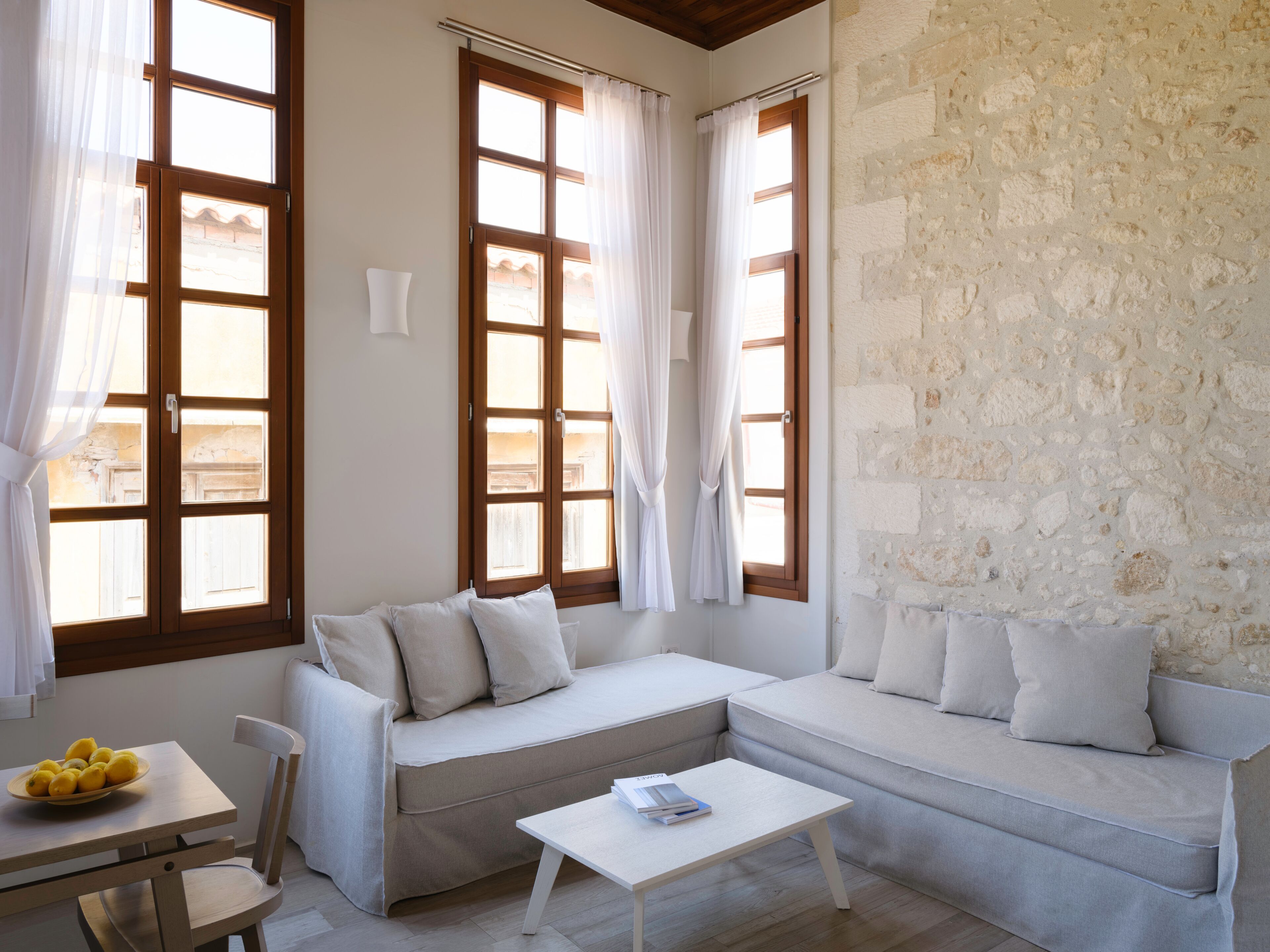 A room with a stone wall, a sofa, a table with a bowl of lemons, and large windows with curtains.