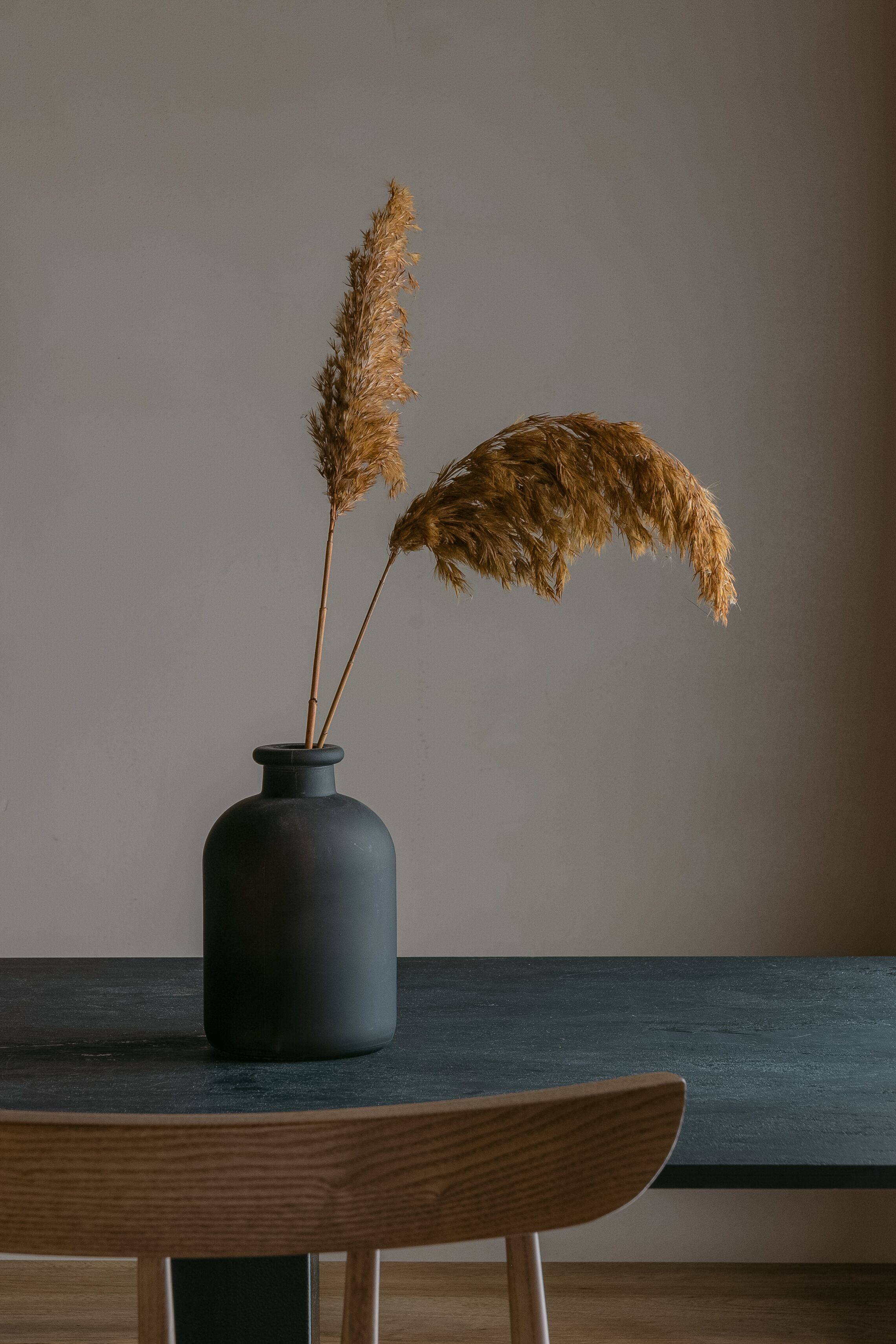 A dark-colored vase with dried pampas grass on a wooden table against a plain wall.