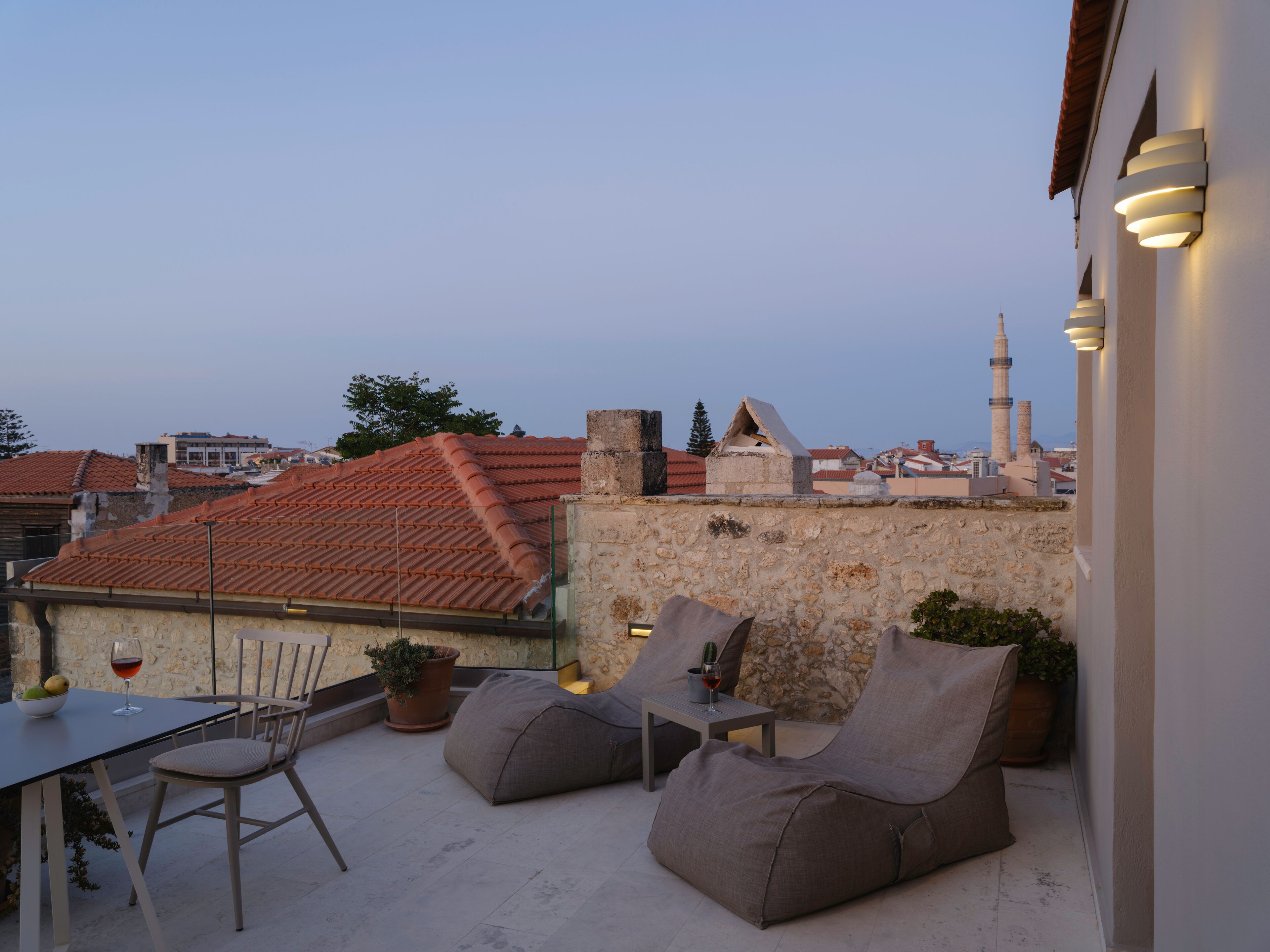 A rooftop terrace with a table, chairs, and bean bags. There are potted plants and a view of buildings and a mosque in the background.