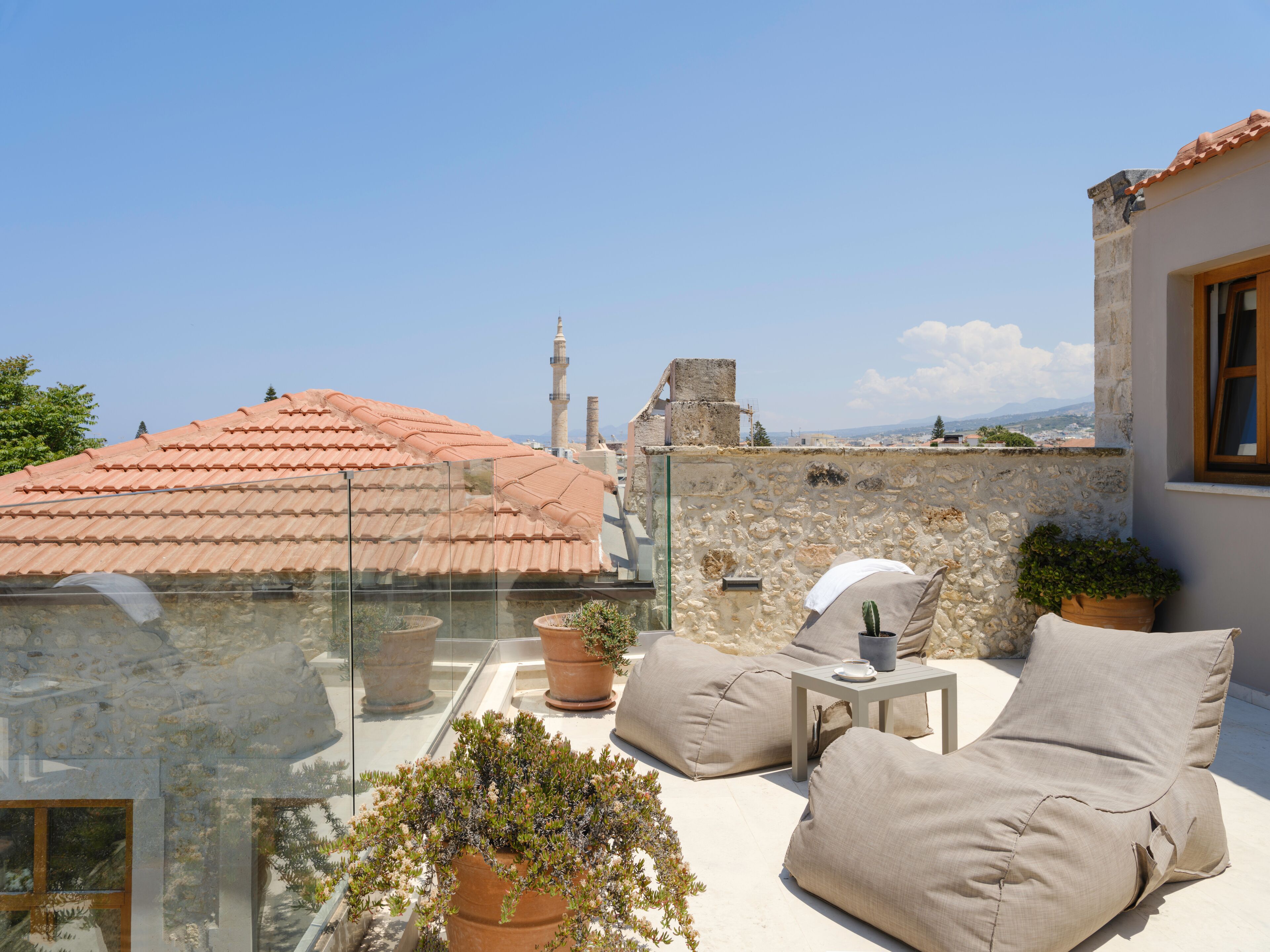Eine Dachterrasse mit Glasgeländer, beigen Kissen, Topfpflanzen und Blick auf Stadtgebäude und eine Moschee.