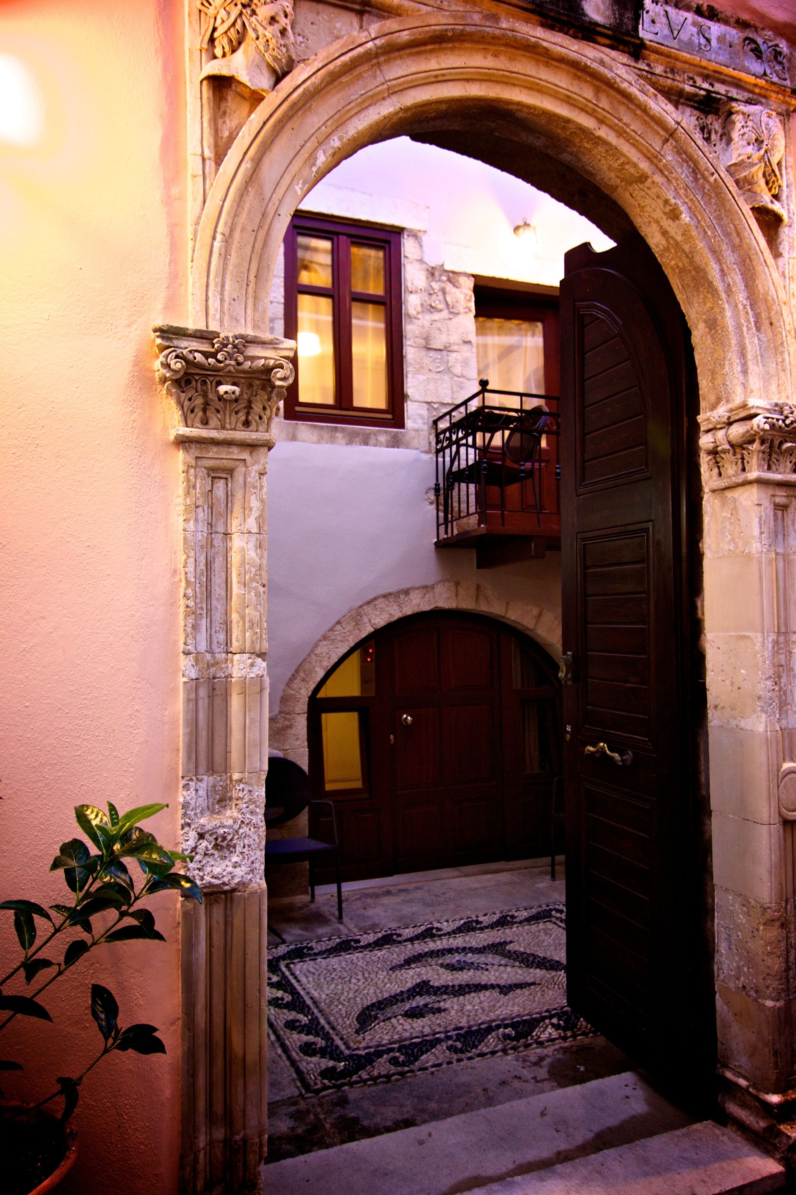 Un arco de entrada con una puerta de madera, un balcón con sillas y una planta en maceta.