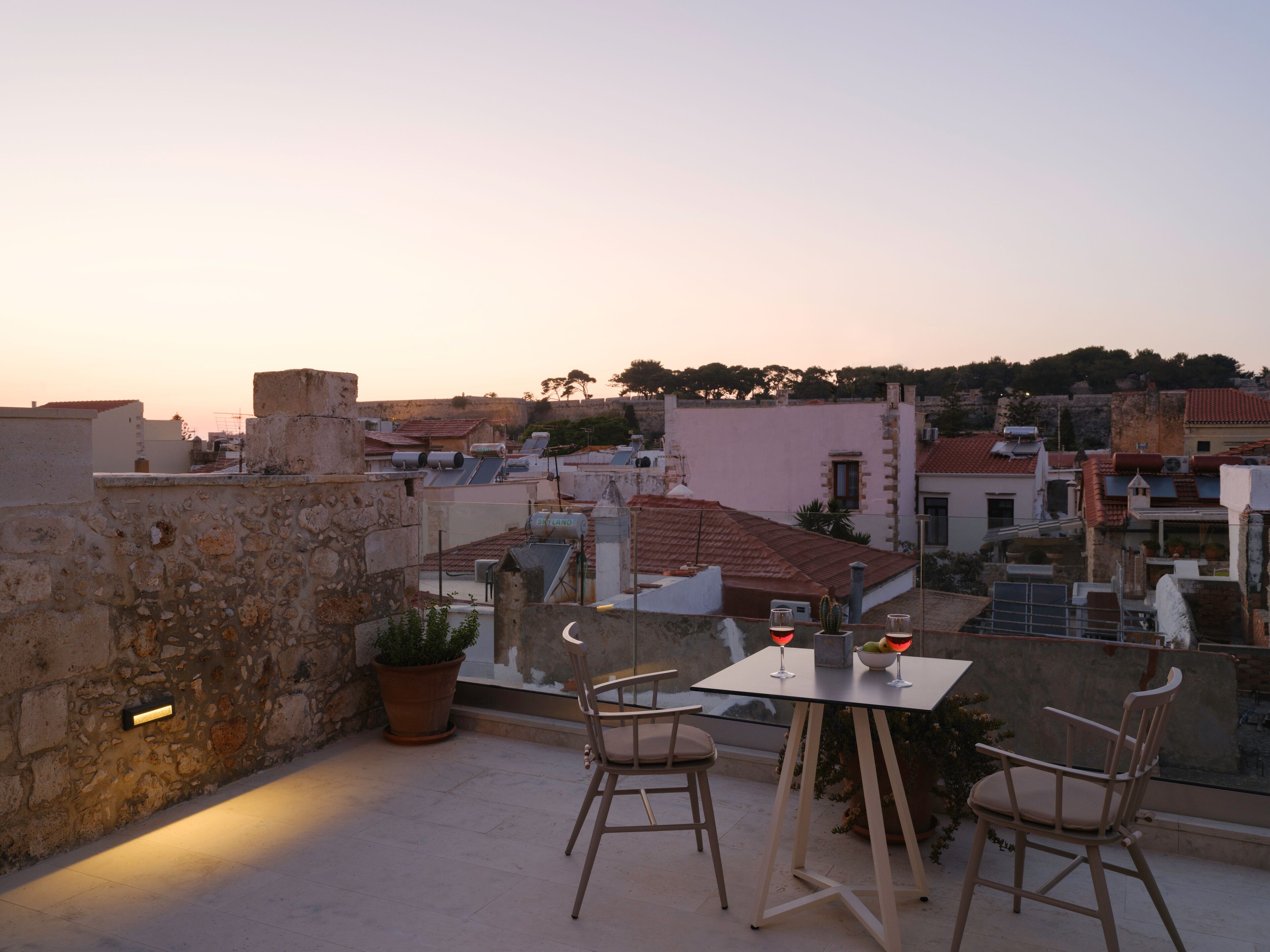 Una terraza con dos sillas y una mesa, con vistas a un paisaje urbano con edificios y un atardecer.