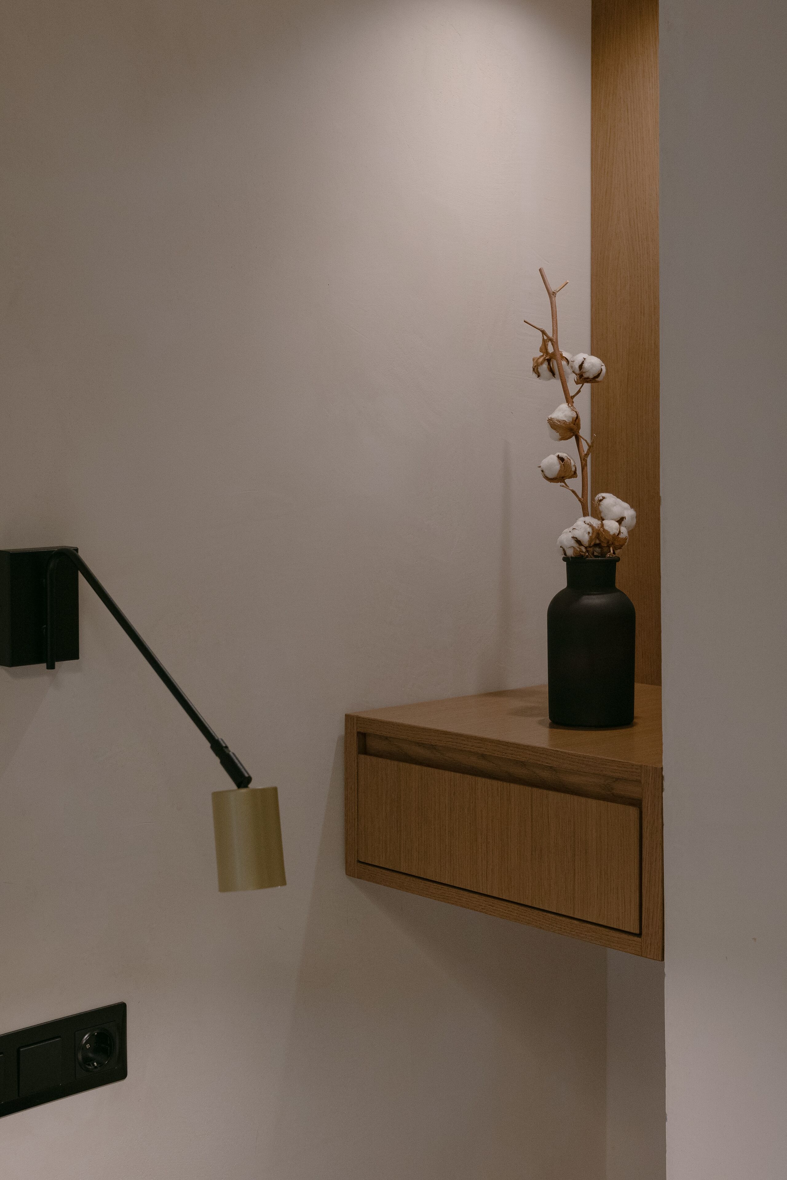A minimalist room with a wooden shelf holding a vase with dried flowers, a wall-mounted lamp, and a wooden cabinet.