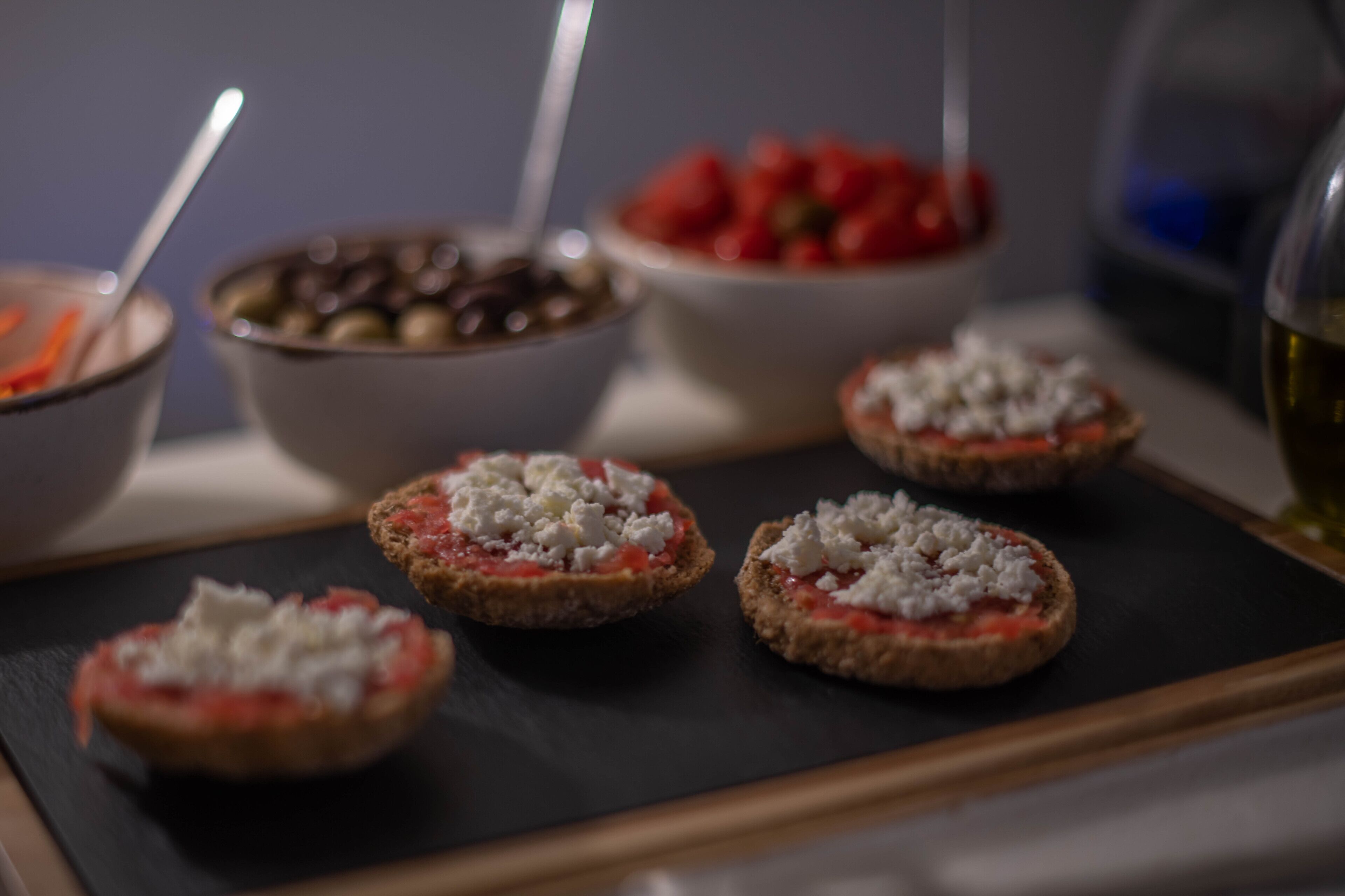 A tray with several open-faced sandwiches topped with cheese and tomato, accompanied by bowls of olives and a bowl of sliced tomatoes.