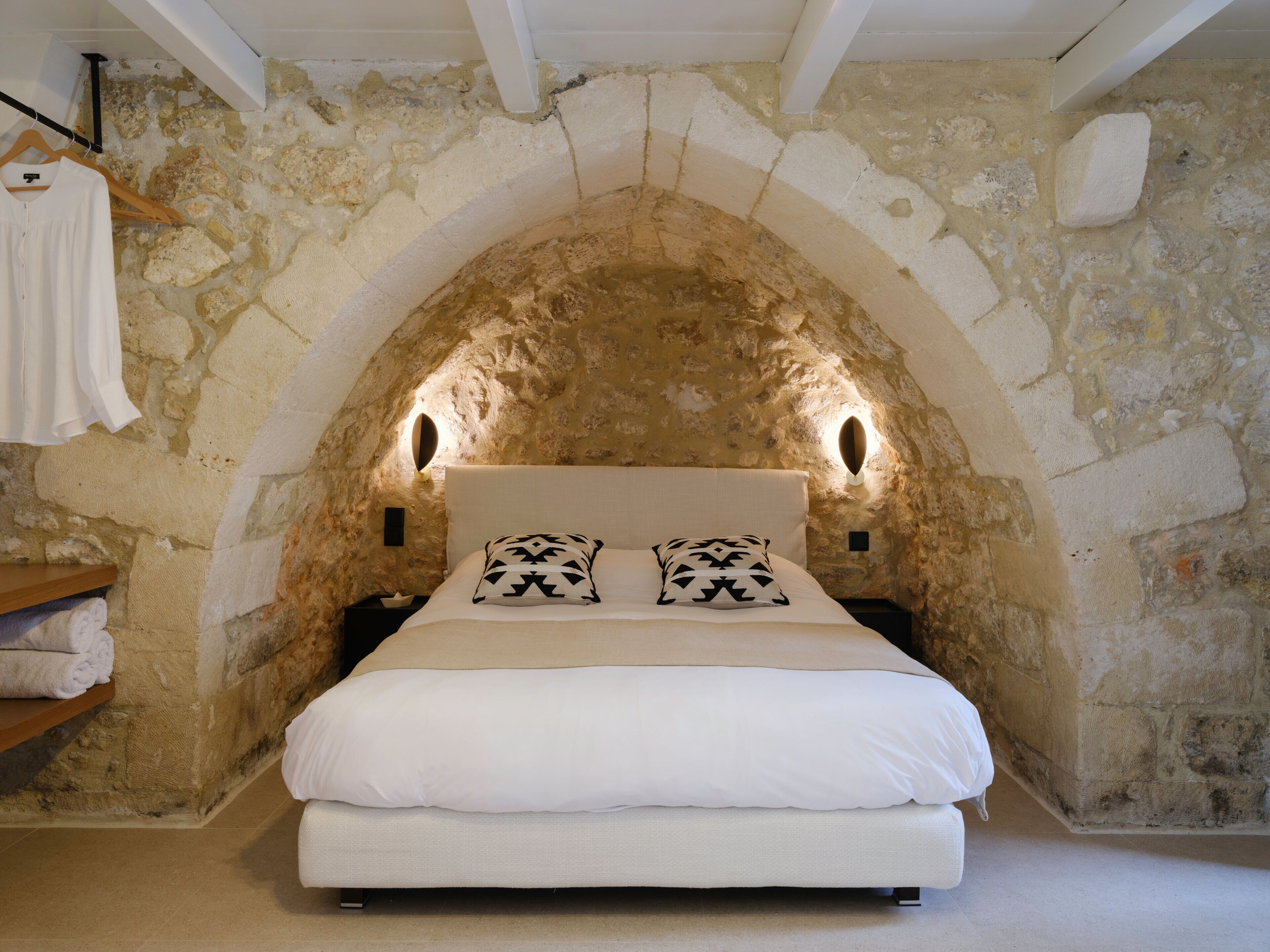 A bedroom with a stone wall arch, a bed with white bedding, two patterned pillows, and a wooden shelf with towels.