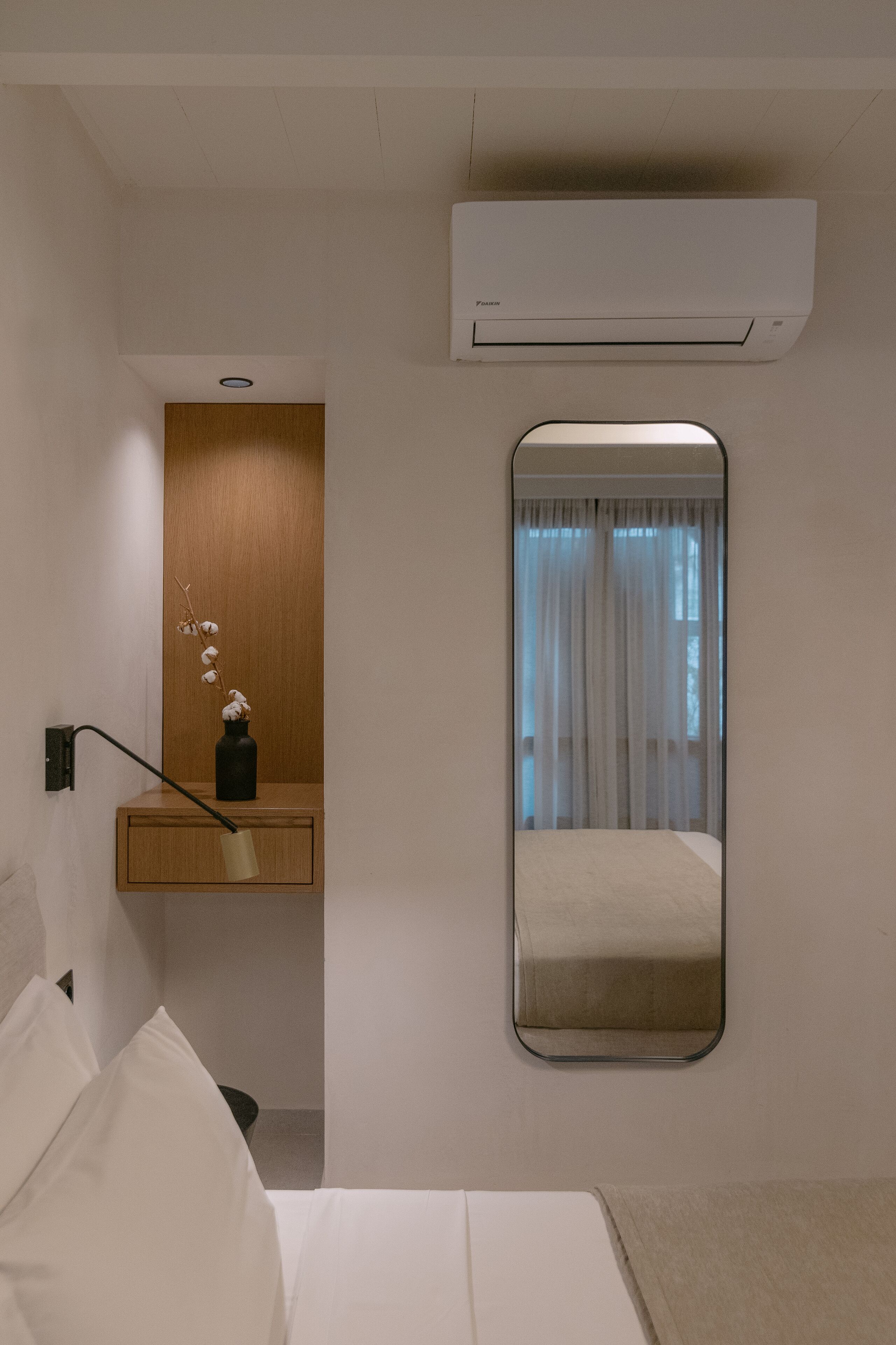 A hotel room with a bed, a bedside table with a lamp, a mirror, and an air conditioning unit.