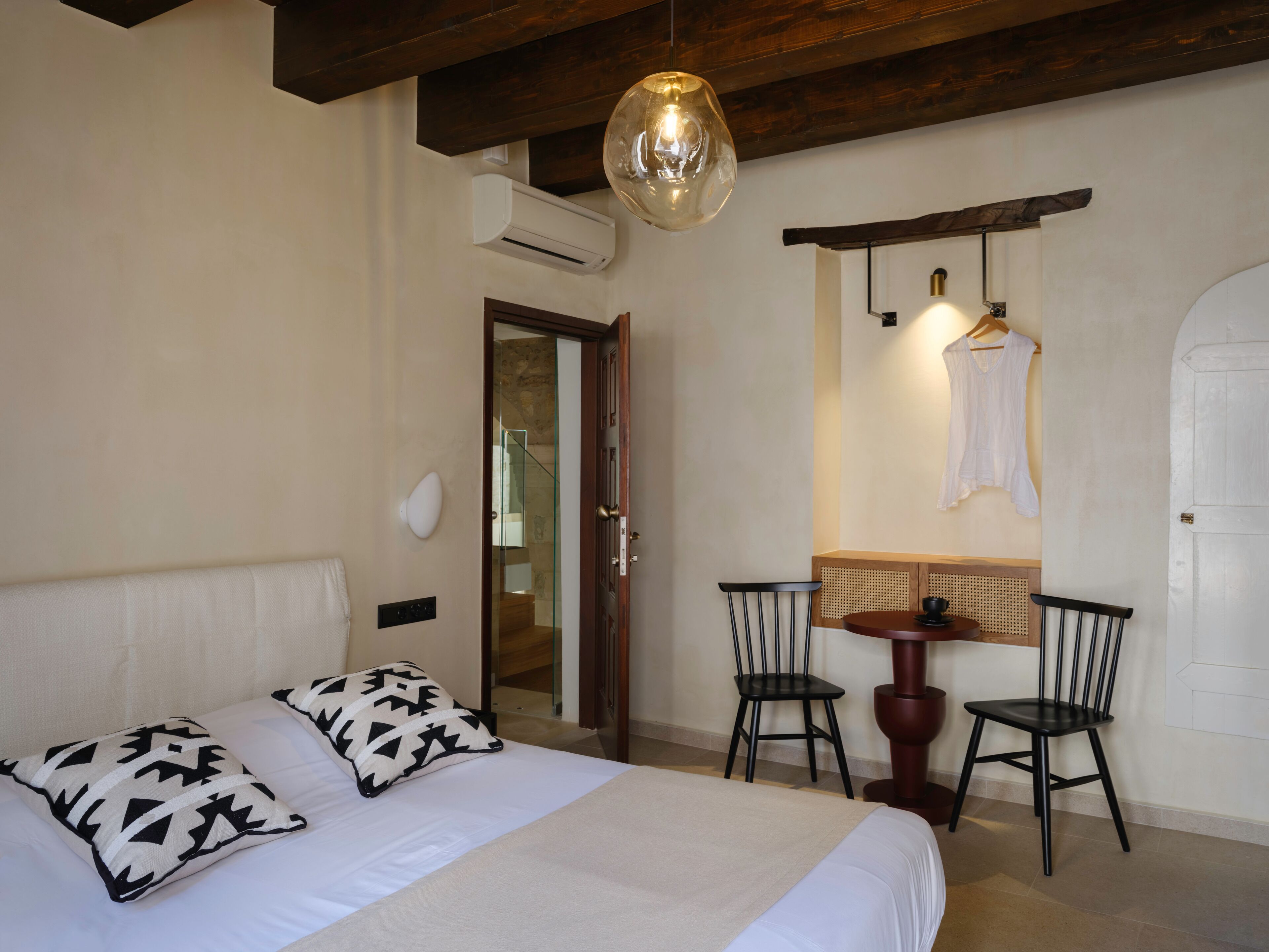 A bedroom with a bed, two chairs, a table, and a hanging light fixture.