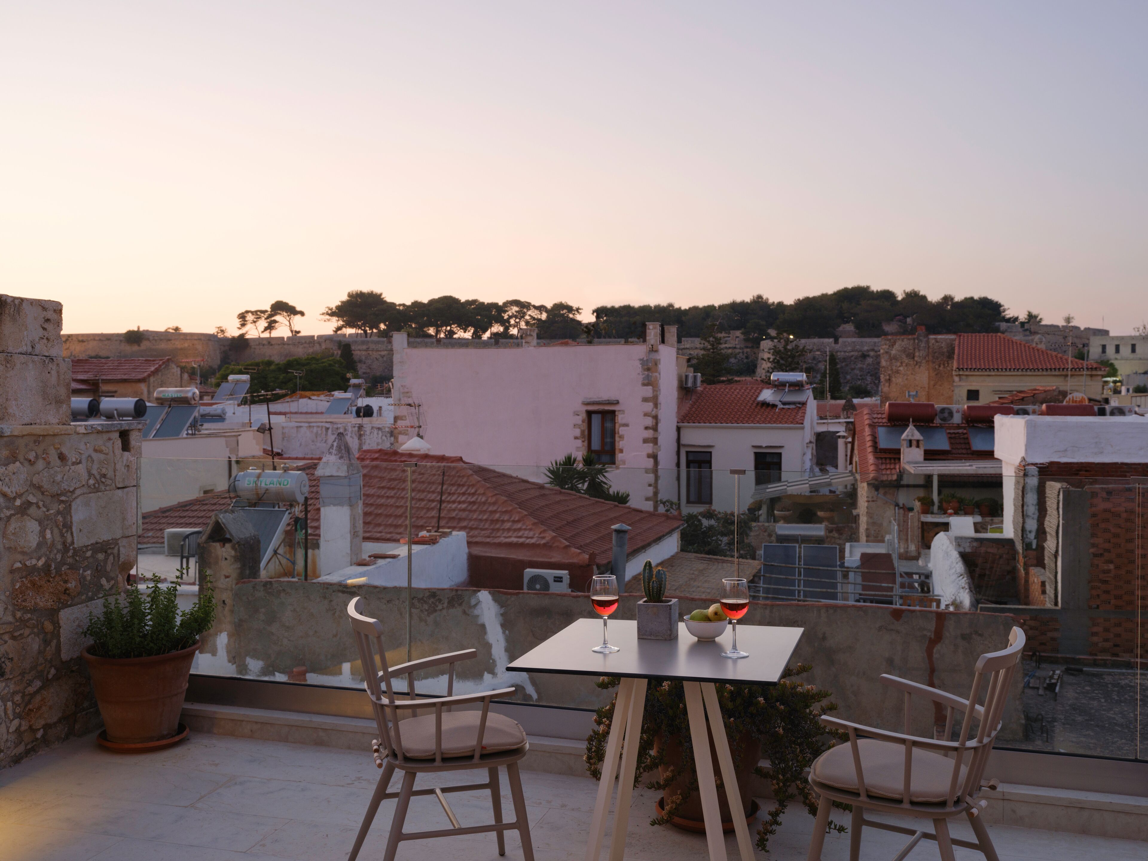 A rooftop terrace with two chairs and a table, two glasses of red wine, and a potted plant.
