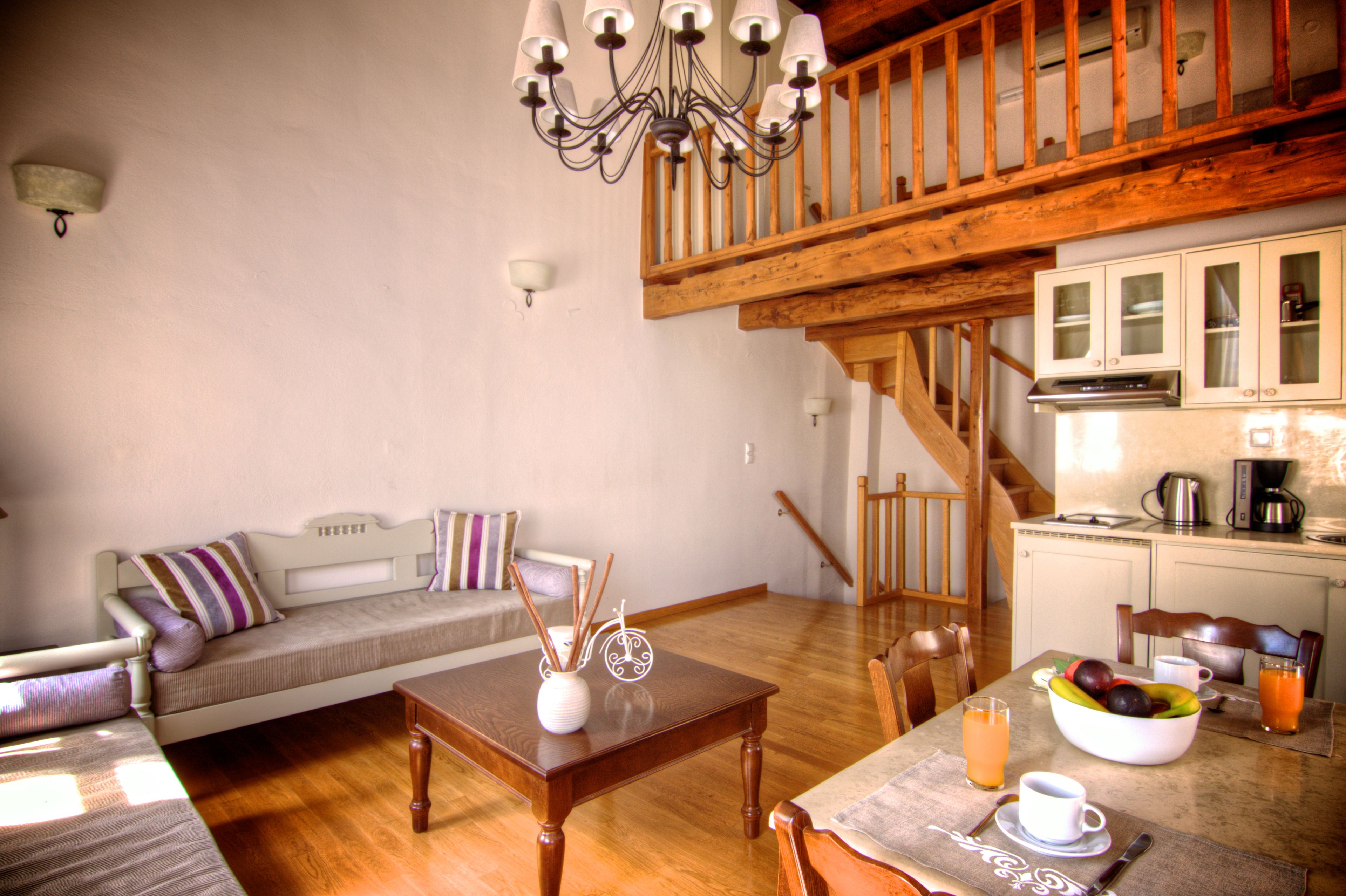 Un salon chaleureux avec un escalier en bois, un coin repas avec une table dressée pour deux personnes, et une cuisine équipée de placards et d’une machine à café.