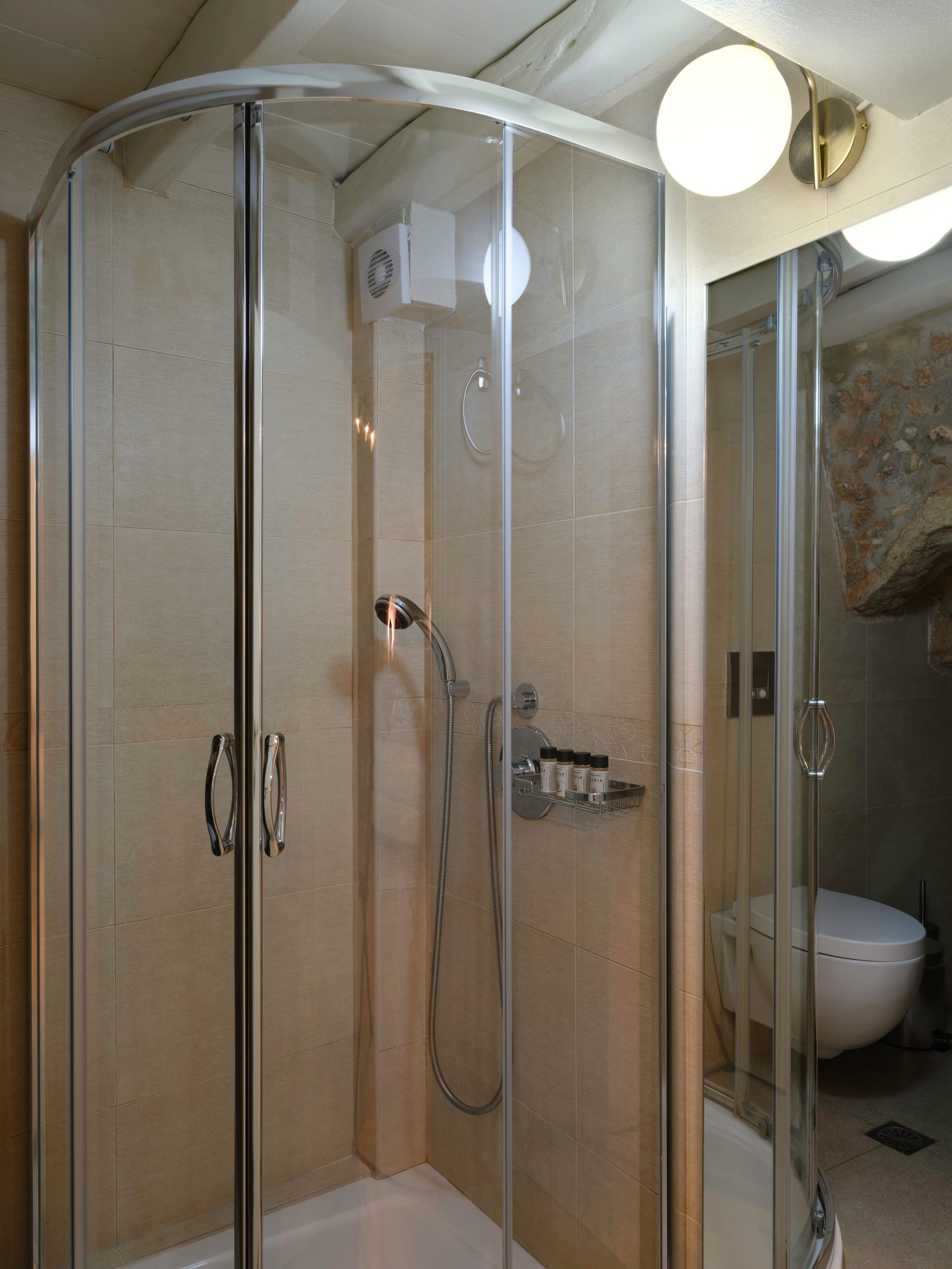 A glass-enclosed shower area with a handheld showerhead, a wall-mounted faucet, and a shelf with toiletries.