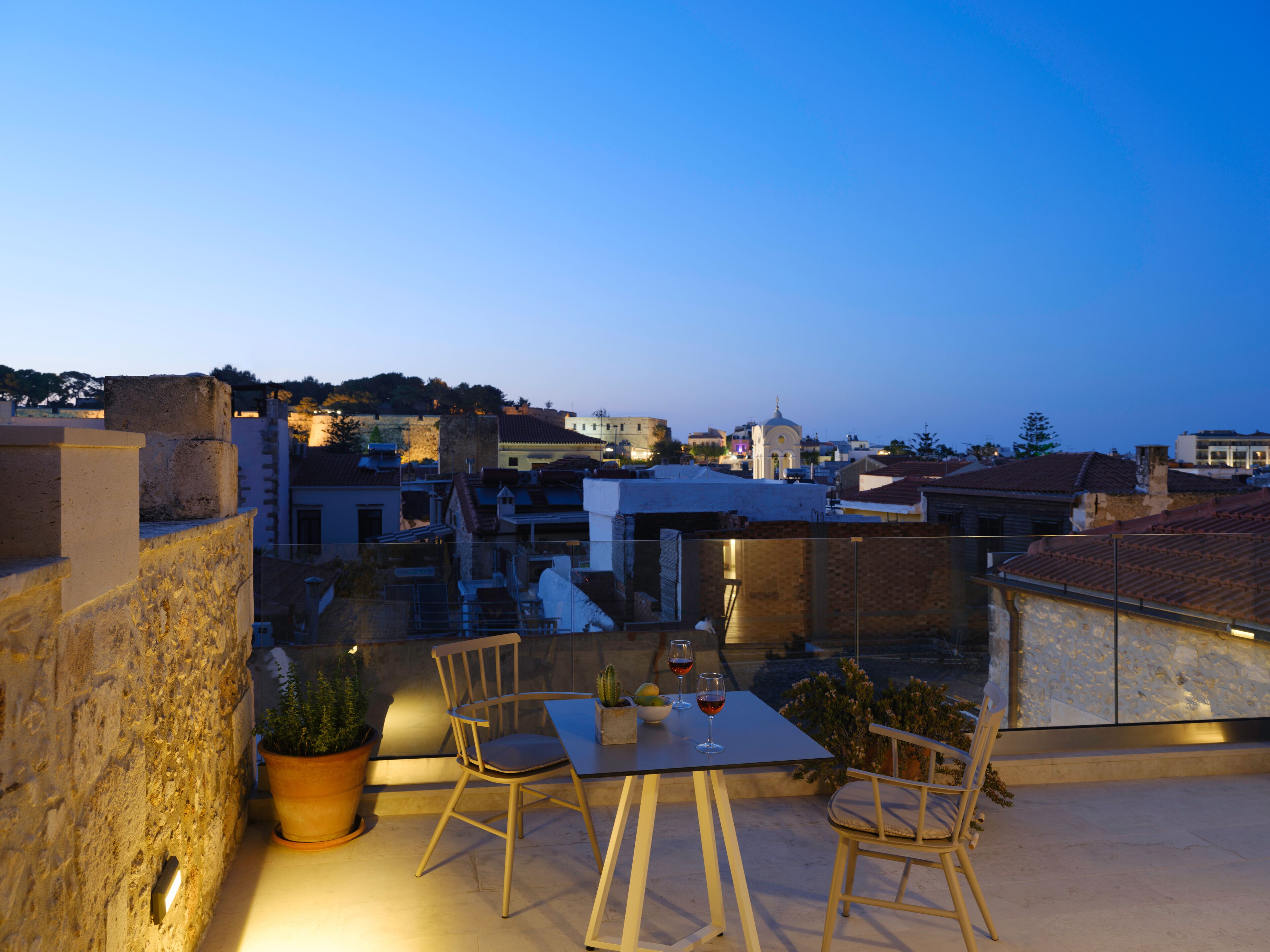 A rooftop terrace with a table and chairs, potted plants, and a view of the cityscape at dusk.