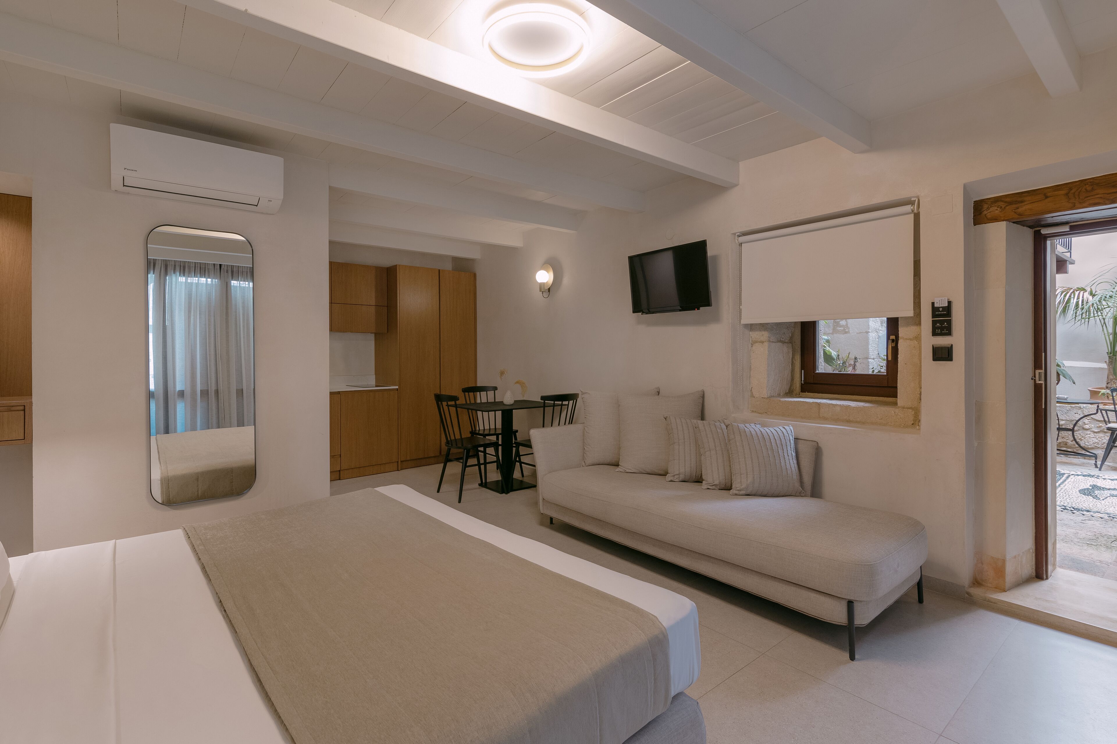A modern hotel room with a bed, a sofa, a dining table with chairs, a TV mounted on the wall, and an air conditioning unit.