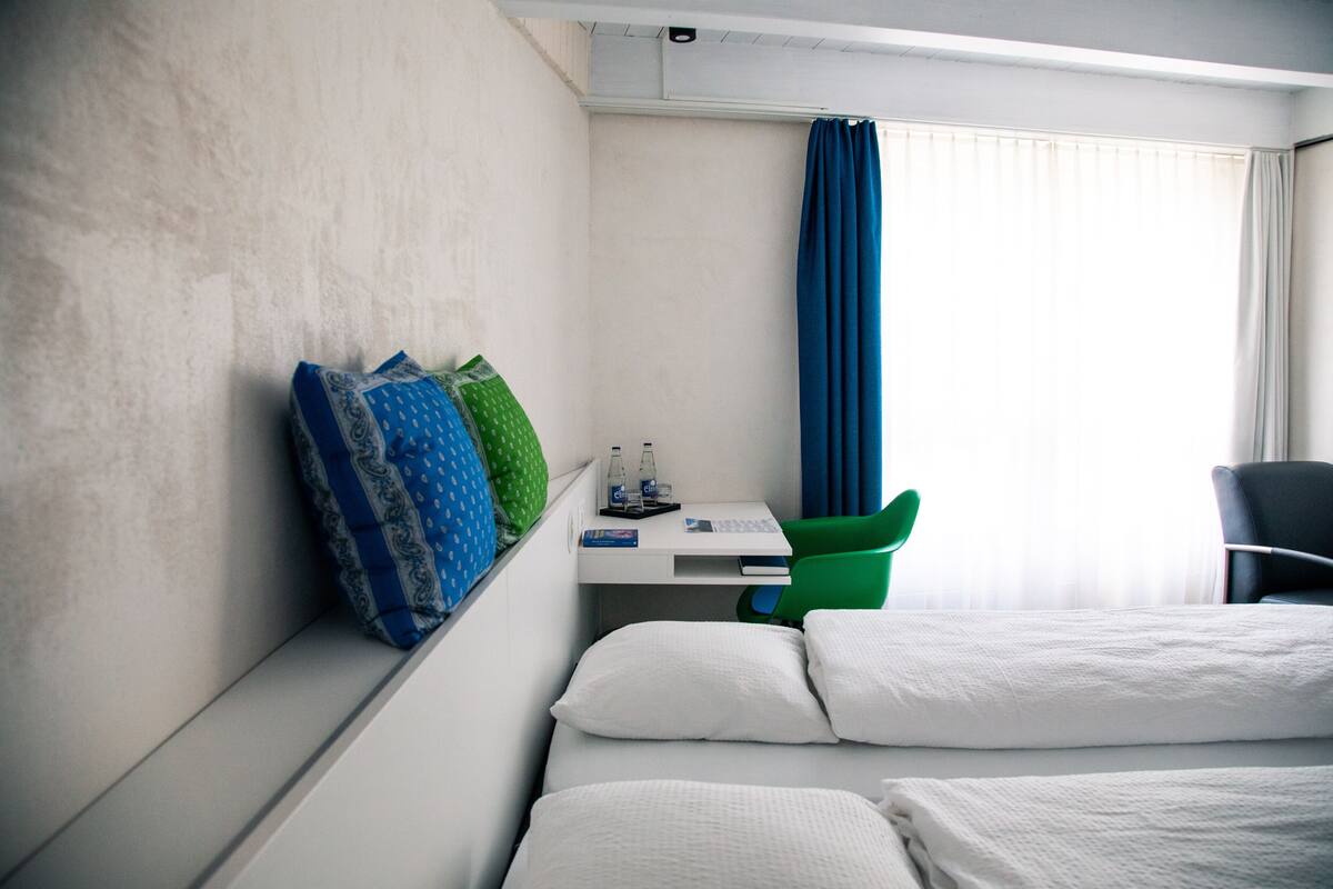 Ein Hotelzimmer mit zwei Betten, einem Schreibtisch mit Stuhl, einem Fenster mit Vorhängen und einer Wand mit Kissen.