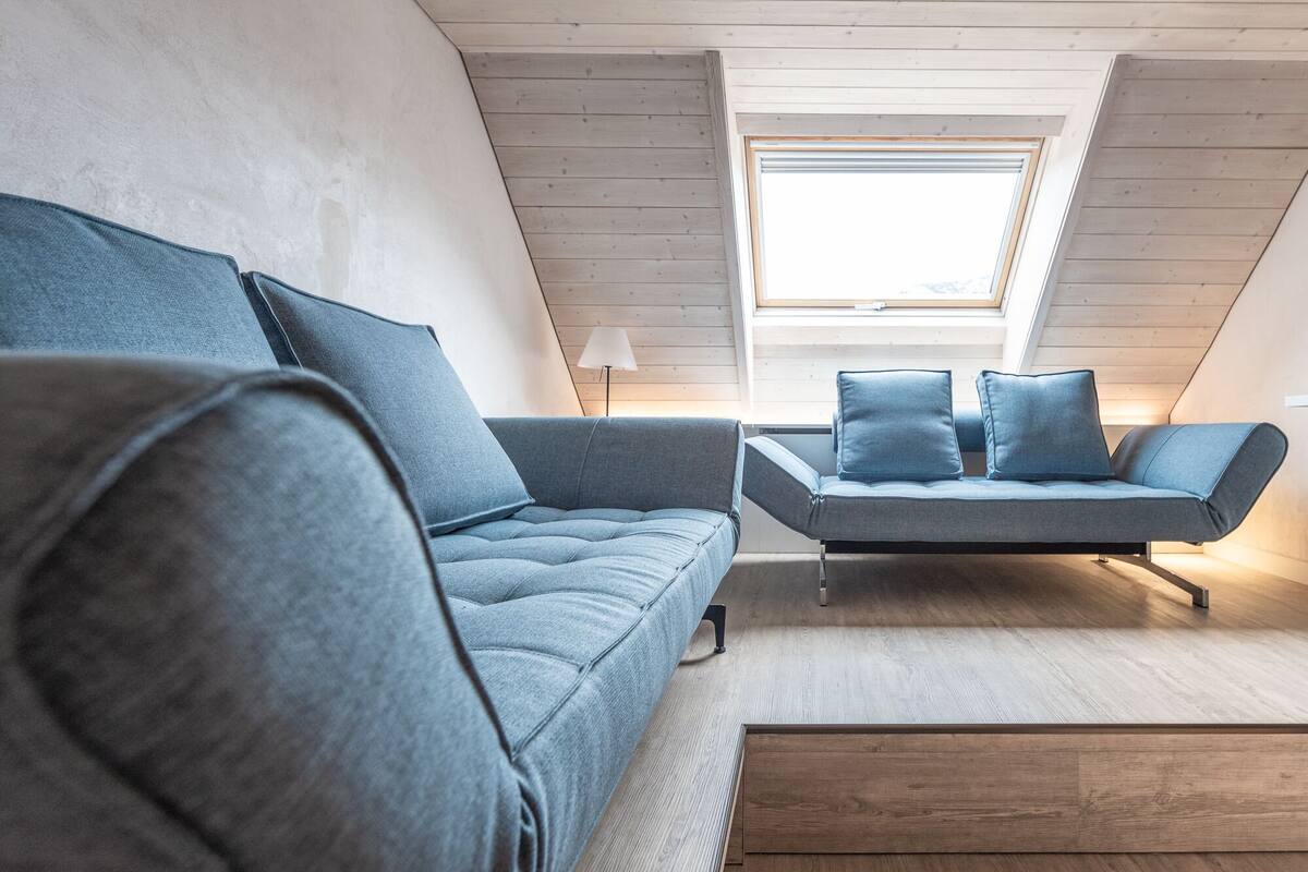 Ein modernes Schlafzimmer im Loft-Stil mit schrägem Dach, Dachfenster und einem grauen Sofa mit Kissen.