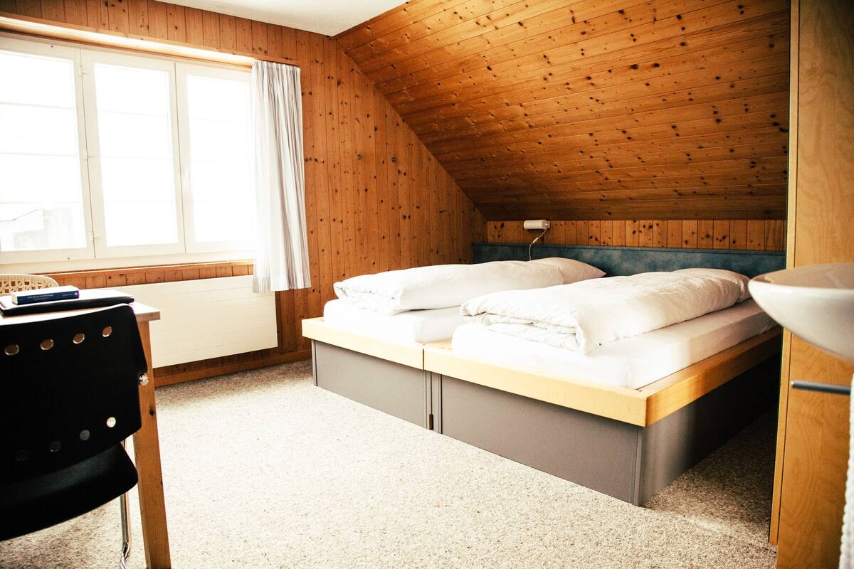 Ein Schlafzimmer mit Holzbalkendecke, einem Bett mit weißen Leinentüchern, einem Schreibtisch mit Stuhl und einem Fenster mit Vorhängen.