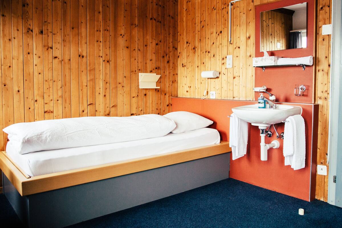 Ein Hotelzimmer mit einer holzgetäfelten Wand, einem Bett mit weißer Bettwäsche, einem Badezimmer mit Waschbecken und Spiegel sowie einem blauen Teppich.