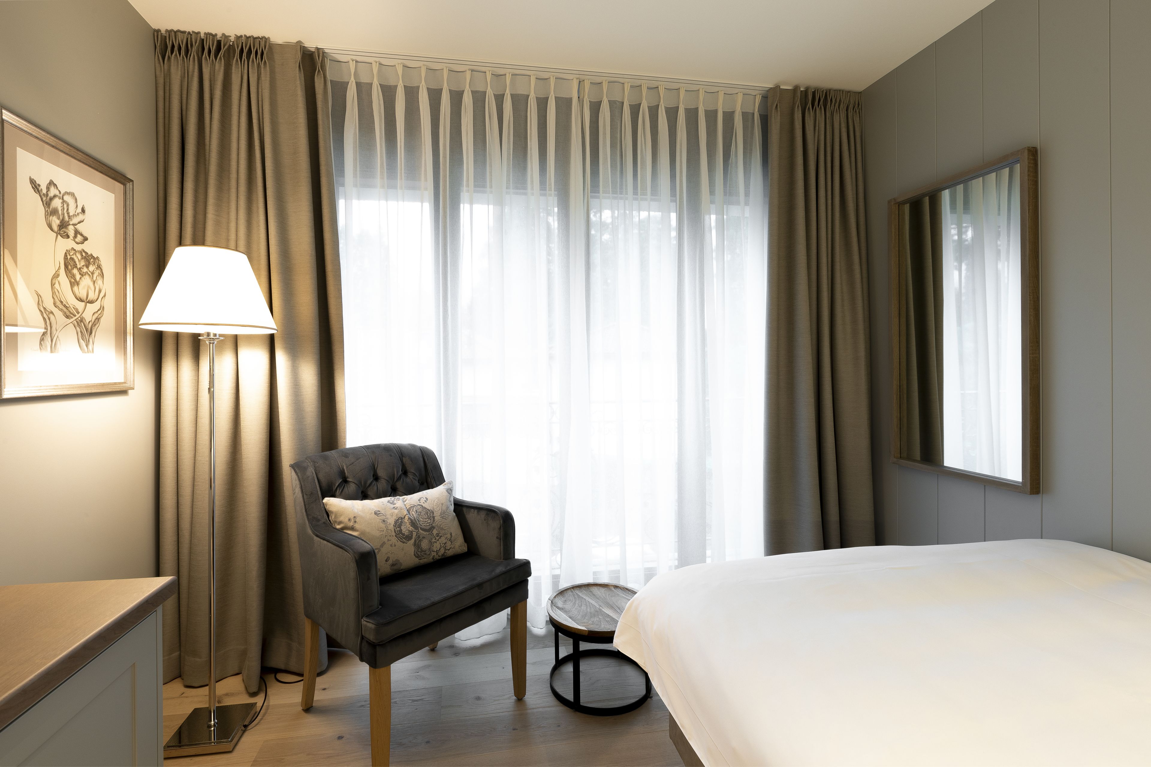 Una habitación de hotel con una cama, una silla, una lámpara, una mesita y una ventana con cortinas.