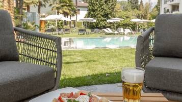 Ein Esstisch mit einer Speiseplatte und einem Bierglas, mit Blick auf einen Pool und eine Außenlounge.