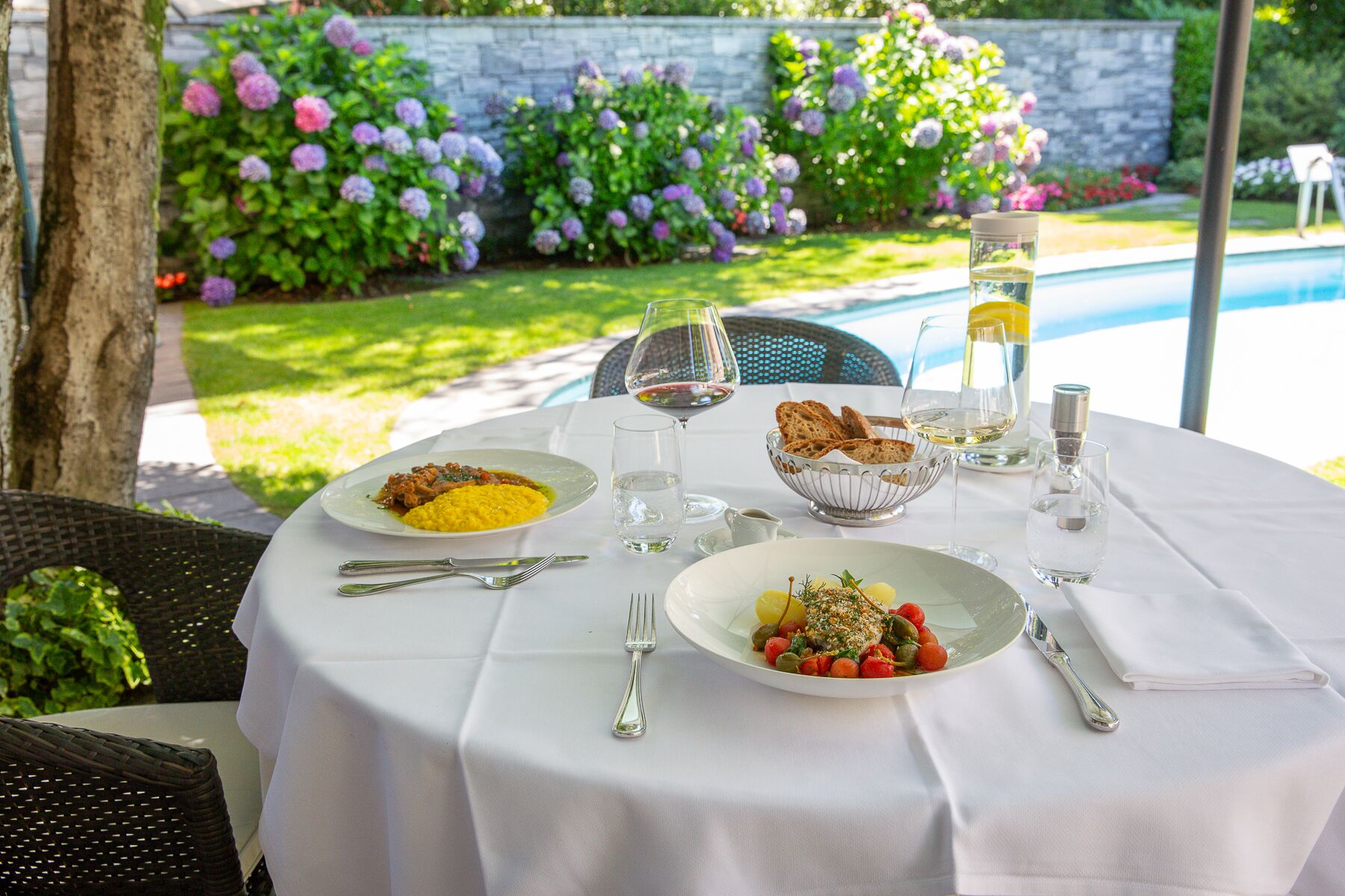 An outdoor dining table set with plates of food, glasses of water and wine, and a basket of bread.